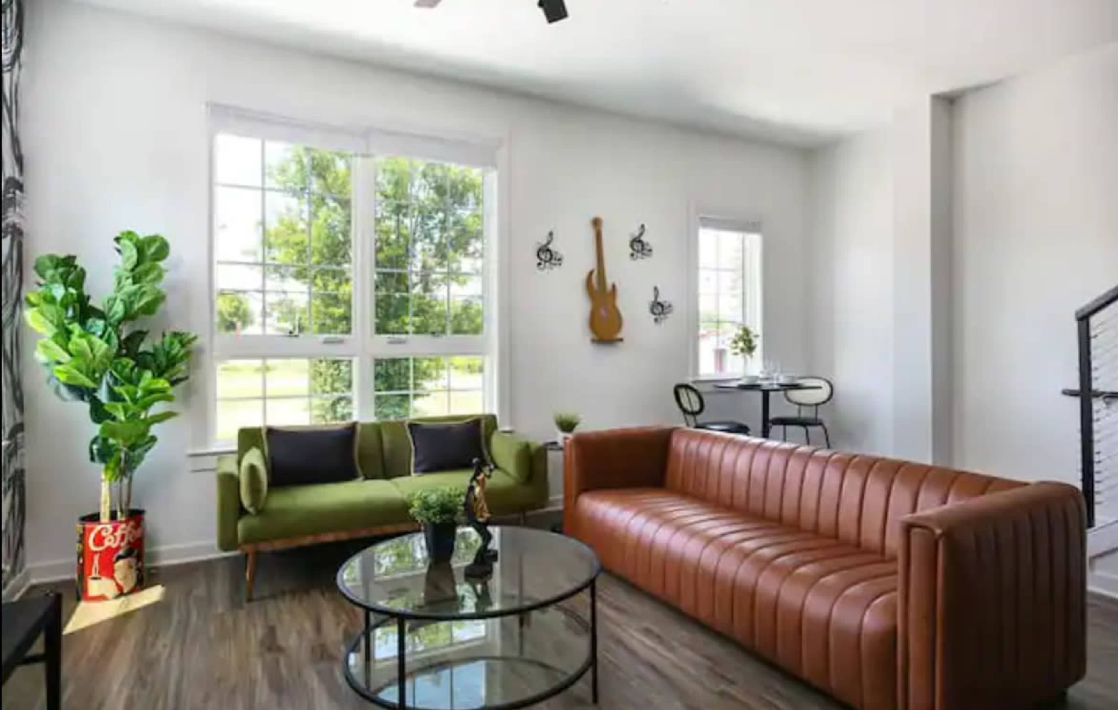 The living room features a green sofa and a brown leather couch arranged around a round glass coffee table, with large windows allowing light to enter the space.