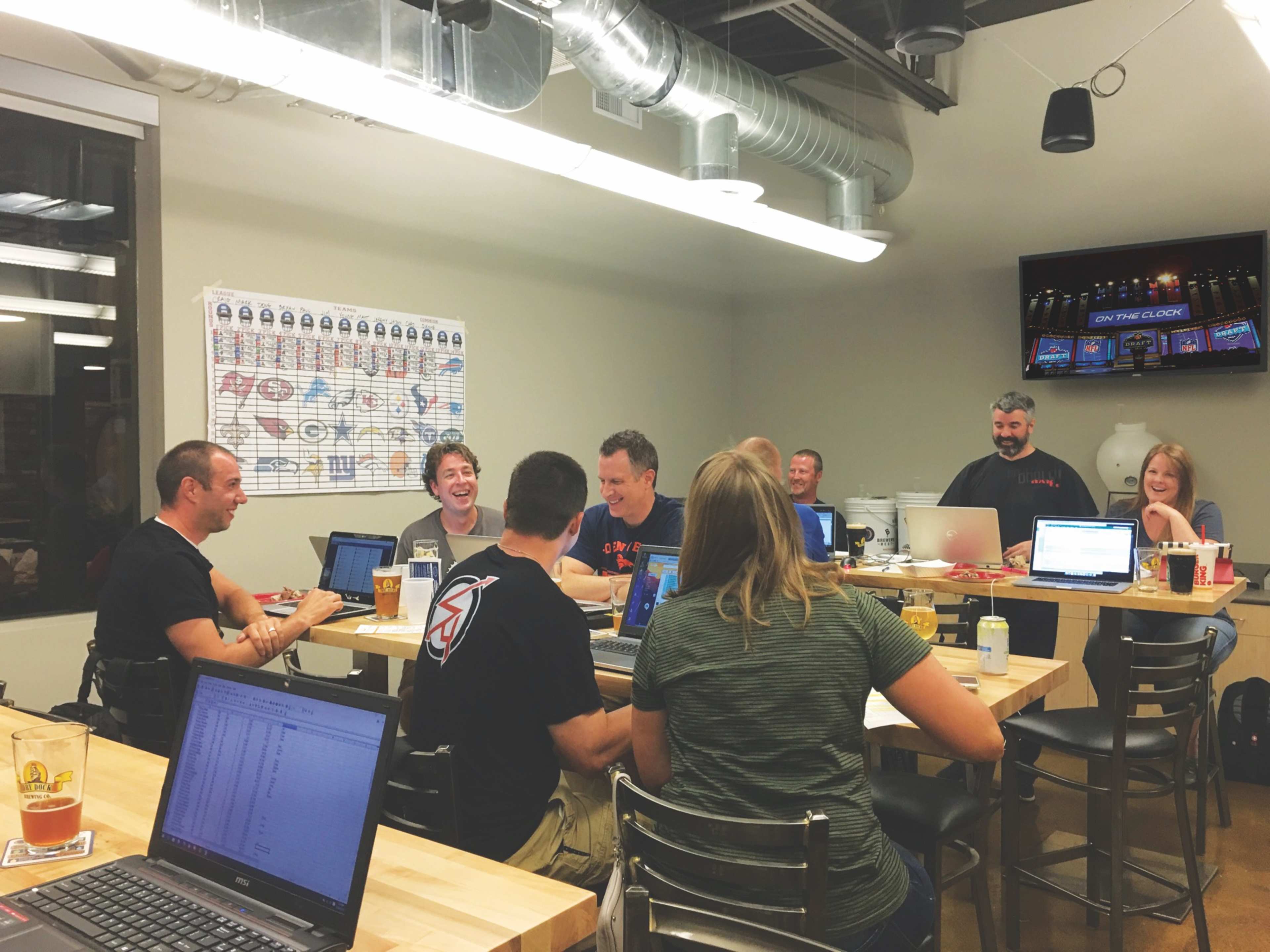A group of people sits around a long table in a well-lit room, working on laptops and engaging in conversation.