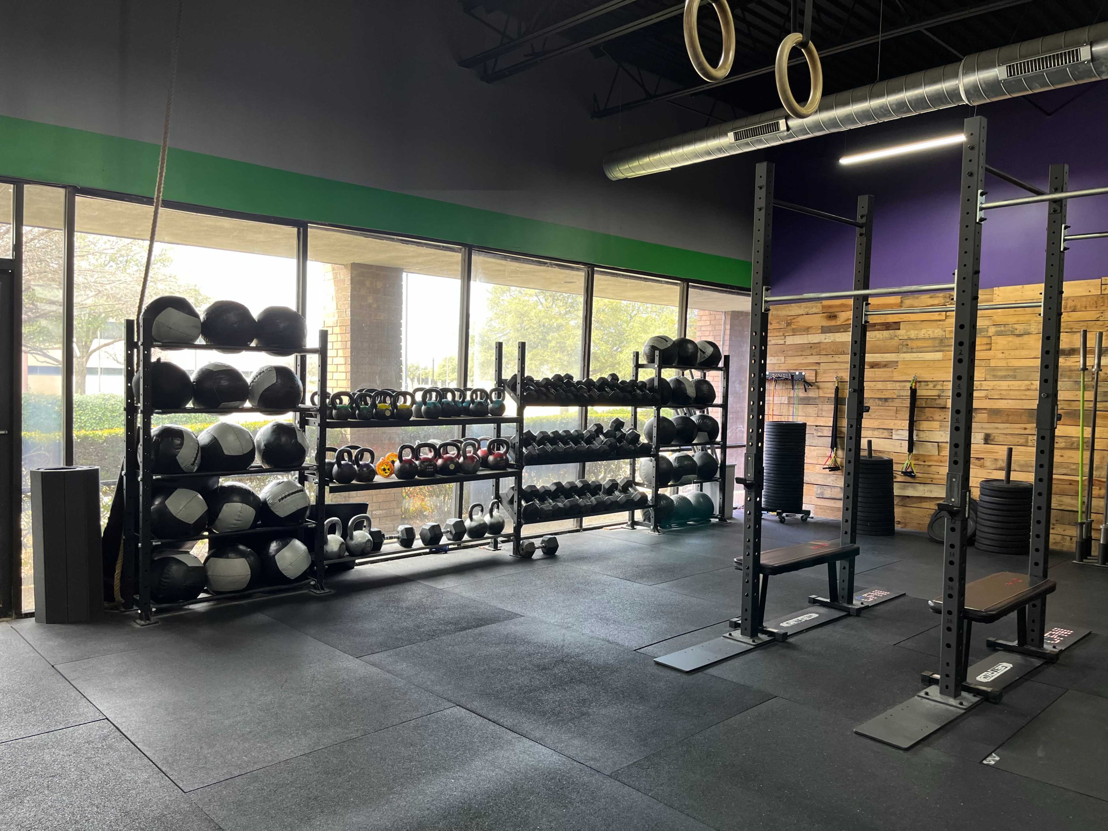 A well-organized gym area with shelves of weights, a squat rack, and various exercise equipment under natural light from large windows.