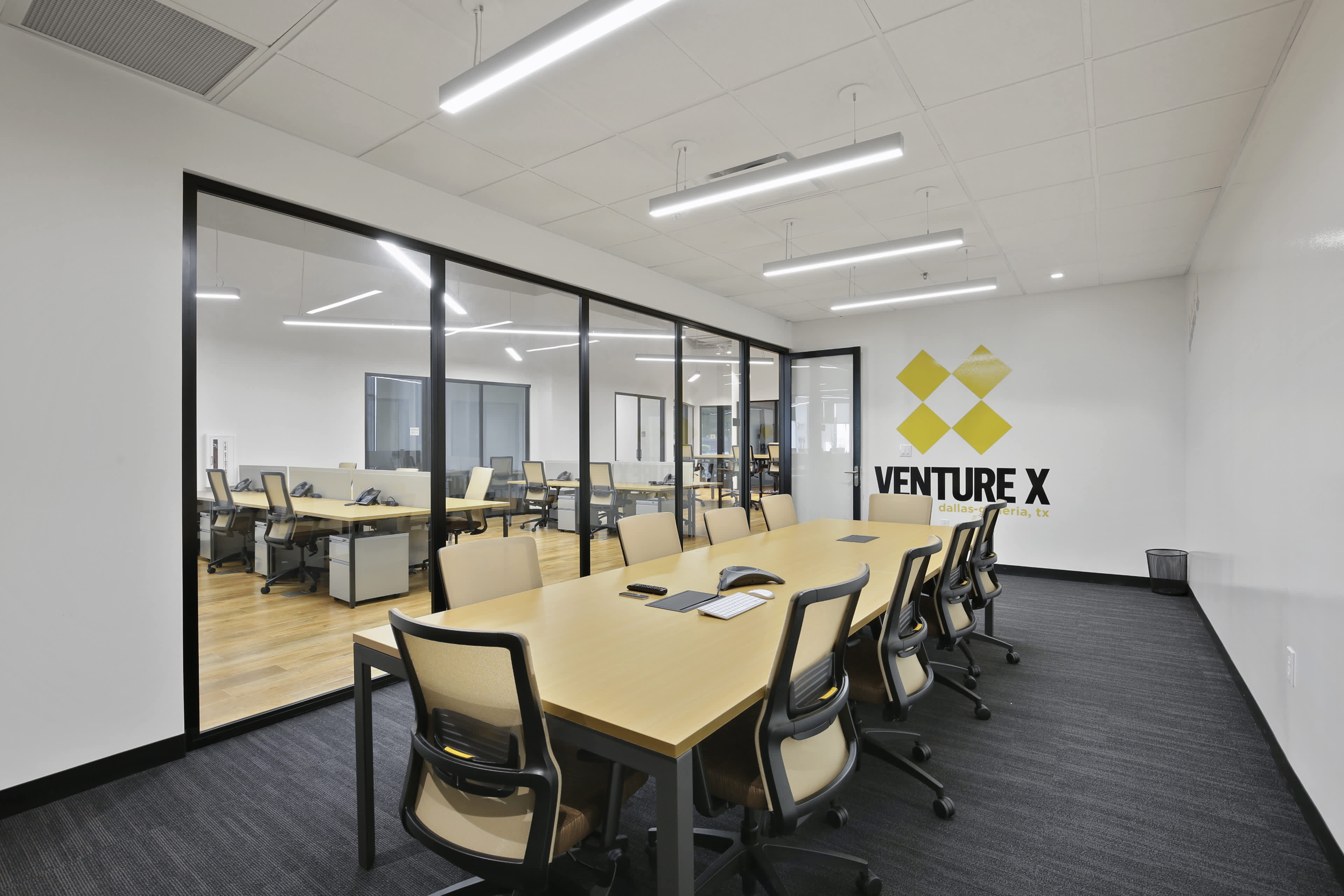 A modern conference room with a large table surrounded by chairs, visible through glass walls, in an office space branded with the "Venture X" logo.