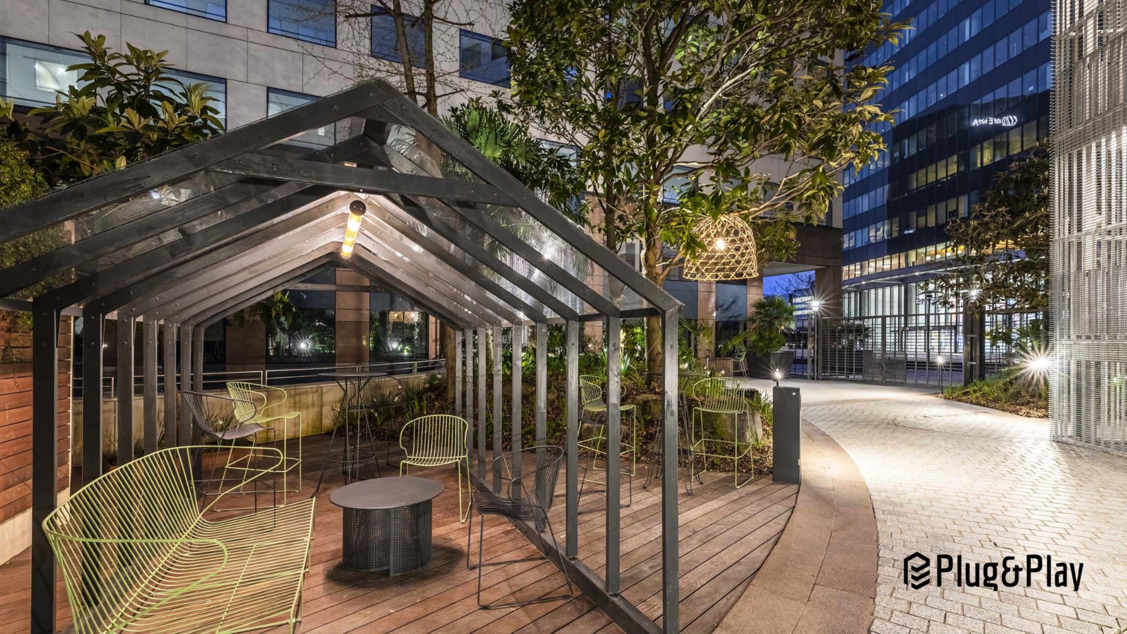 The image features a modern outdoor seating area with a metal frame pergola and stylish chairs surrounded by greenery and contemporary buildings.