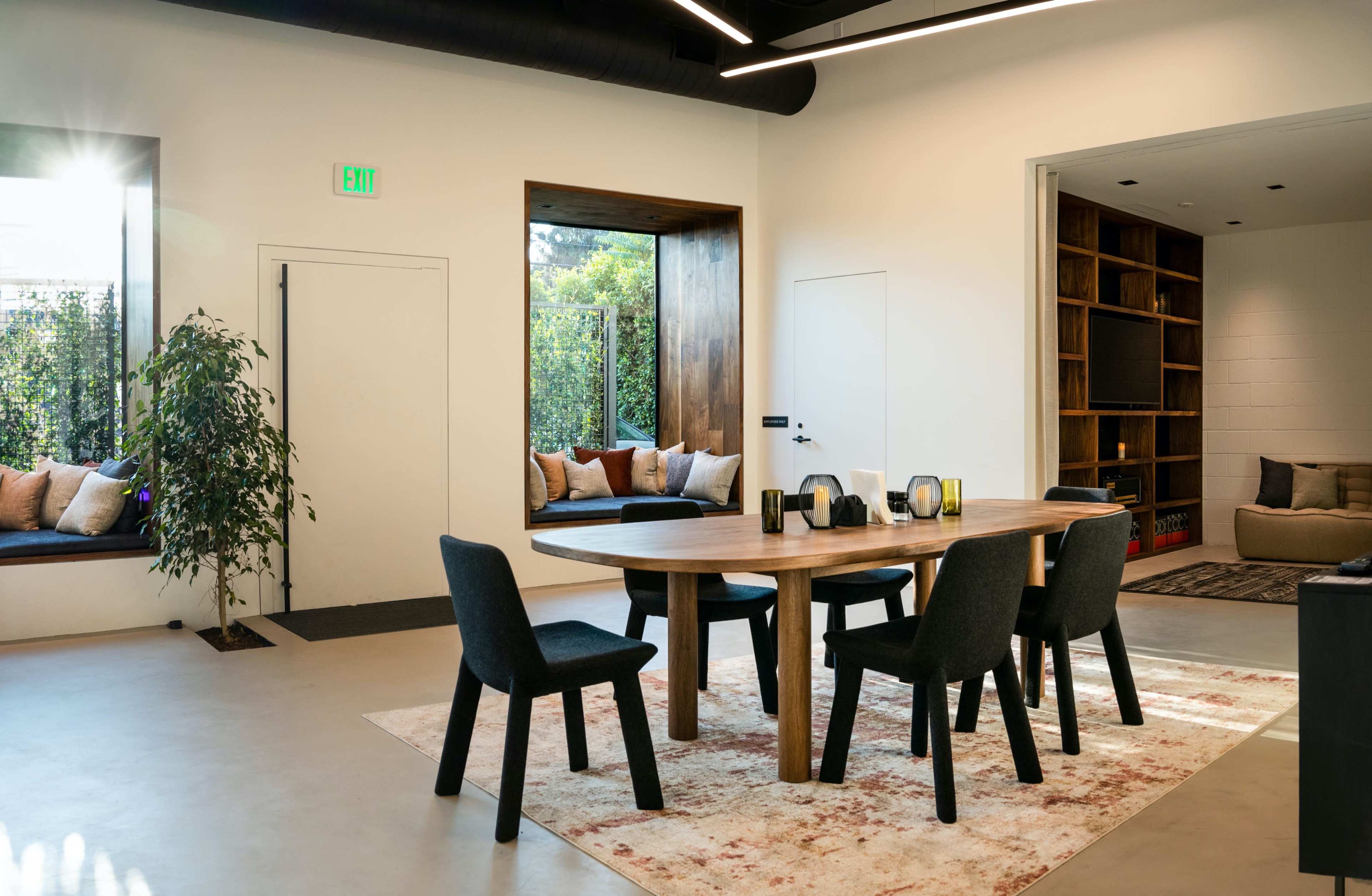 A modern dining area features a wooden table surrounded by black chairs, with built-in benches and greenery visible through large windows.