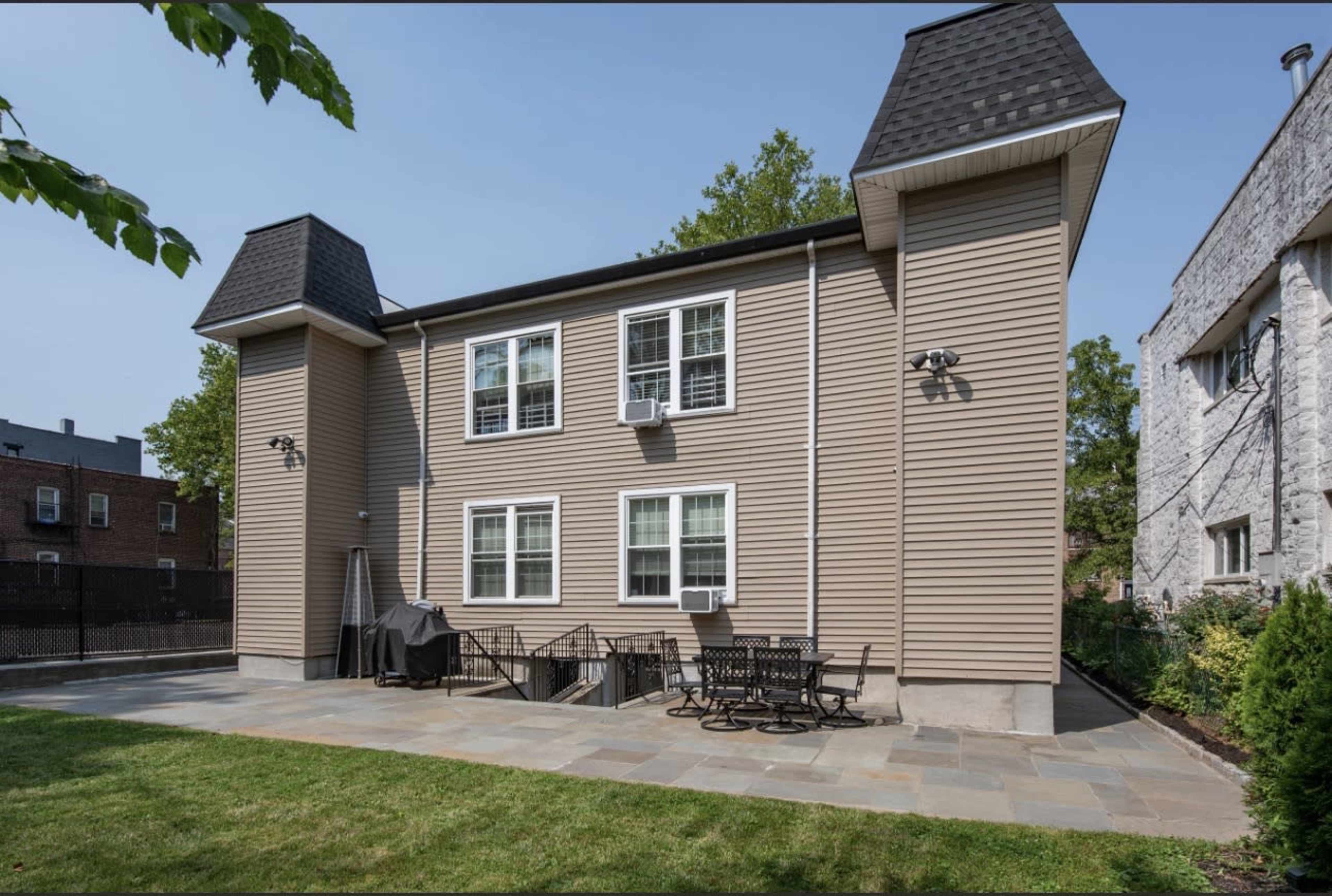 The image shows the exterior of a two-story house with a grey and beige facade, featuring a backyard patio area with stone flooring and patio furniture.