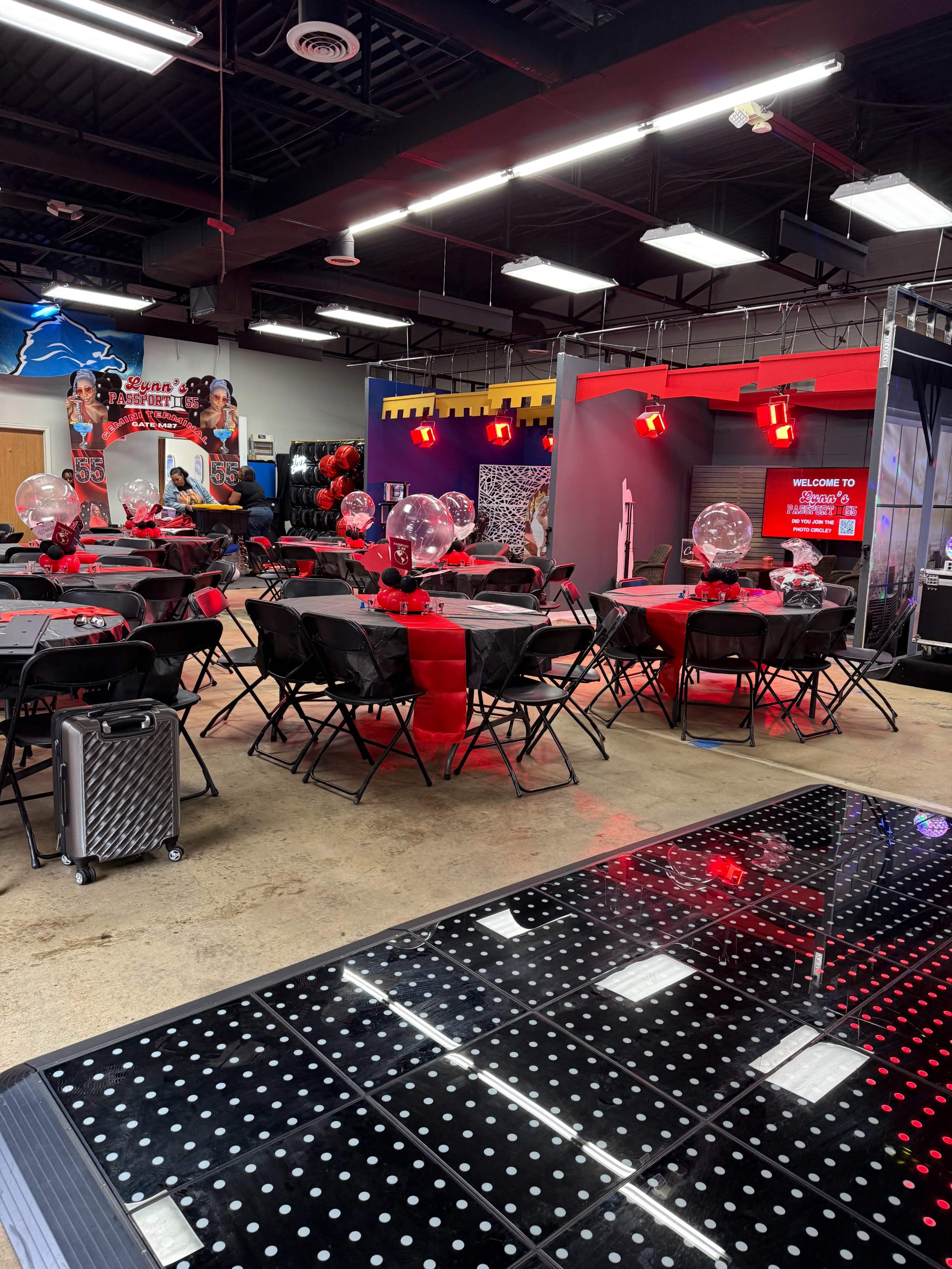 The image shows an indoor event space with decorated tables set up in black and red, featuring balloons and a welcome sign in the background.