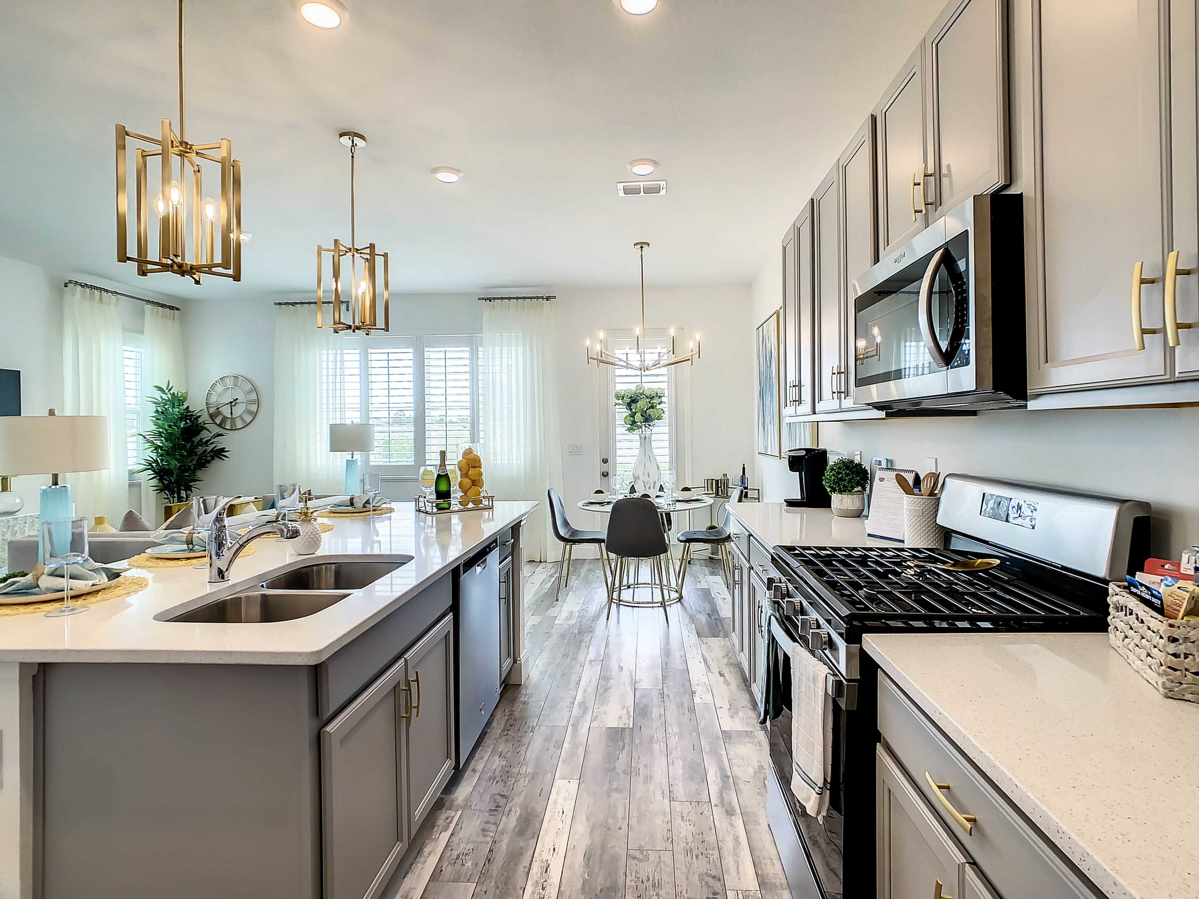 The image shows a modern kitchen featuring gray cabinets, stainless steel appliances, and a dining area with a glass table and stylish light fixtures.