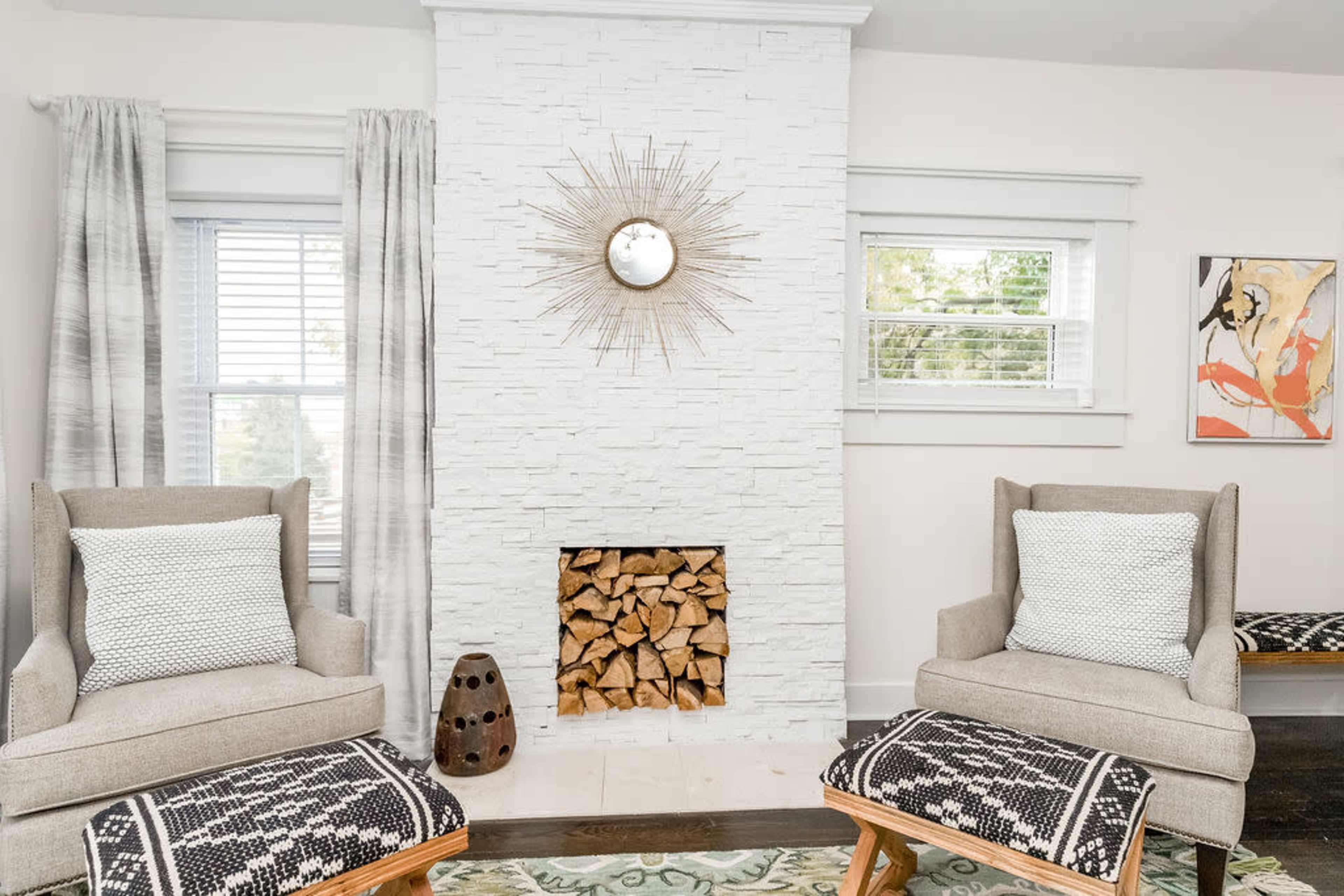 A cozy living room featuring two armchairs with patterned cushions, a stone fireplace with stacked wood, and decorative elements including a wall mirror and artwork.