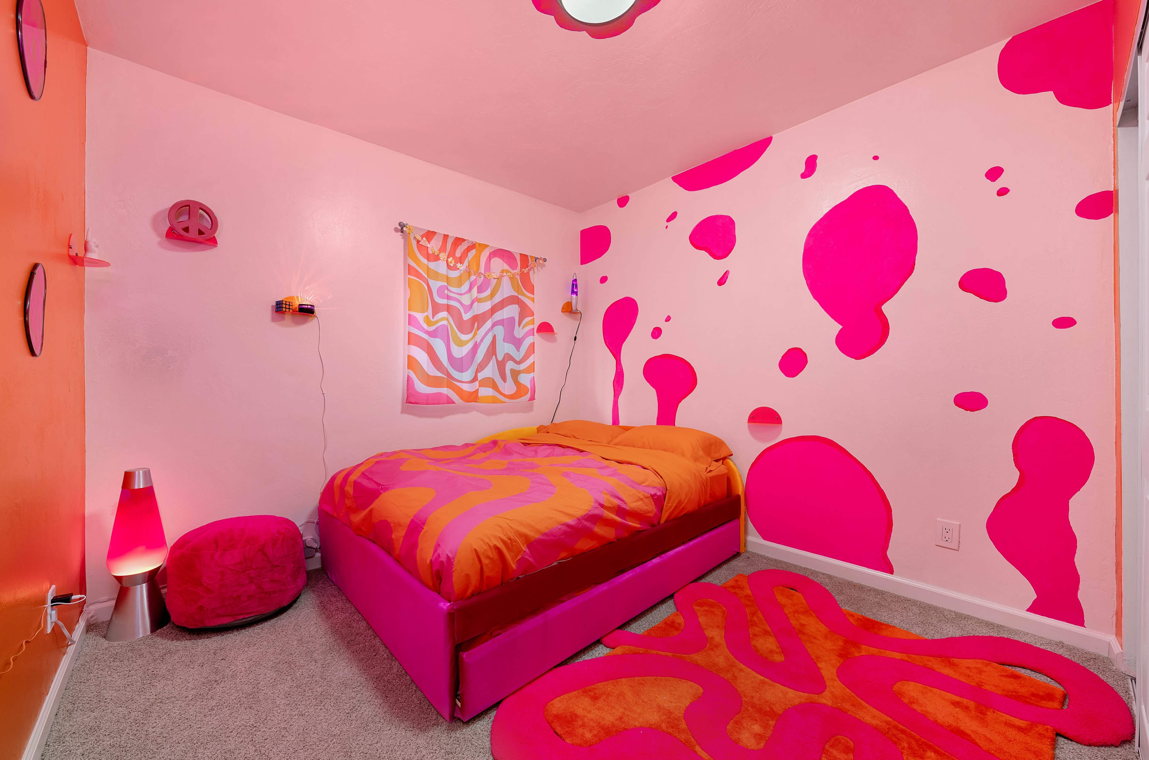 A brightly colored bedroom with pink and orange walls, a bed with an orange comforter, and abstract patterns on the walls and floor.