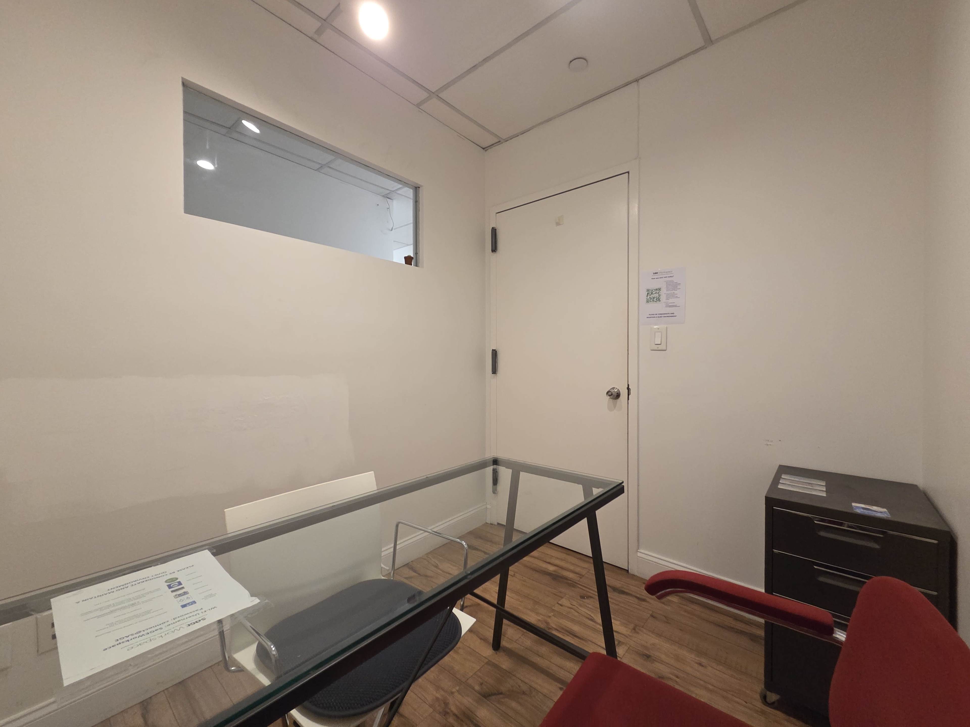 The image shows a small office space with a glass table, a chair, a filing cabinet, and a door, along with a window in the wall to the adjacent area.