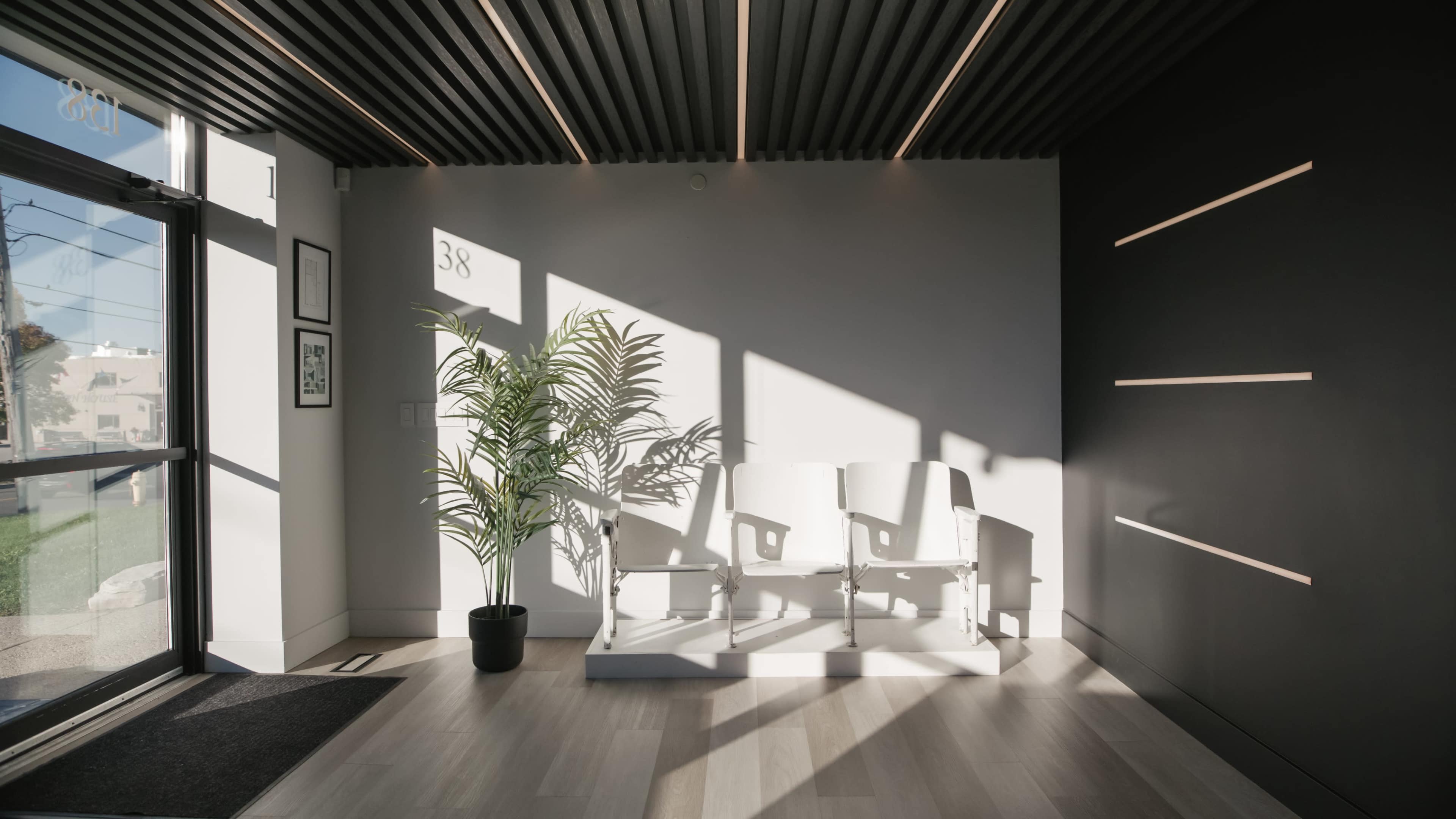 A well-lit waiting area features three white chairs beside a potted plant, with sunlight casting shadows on the floor.