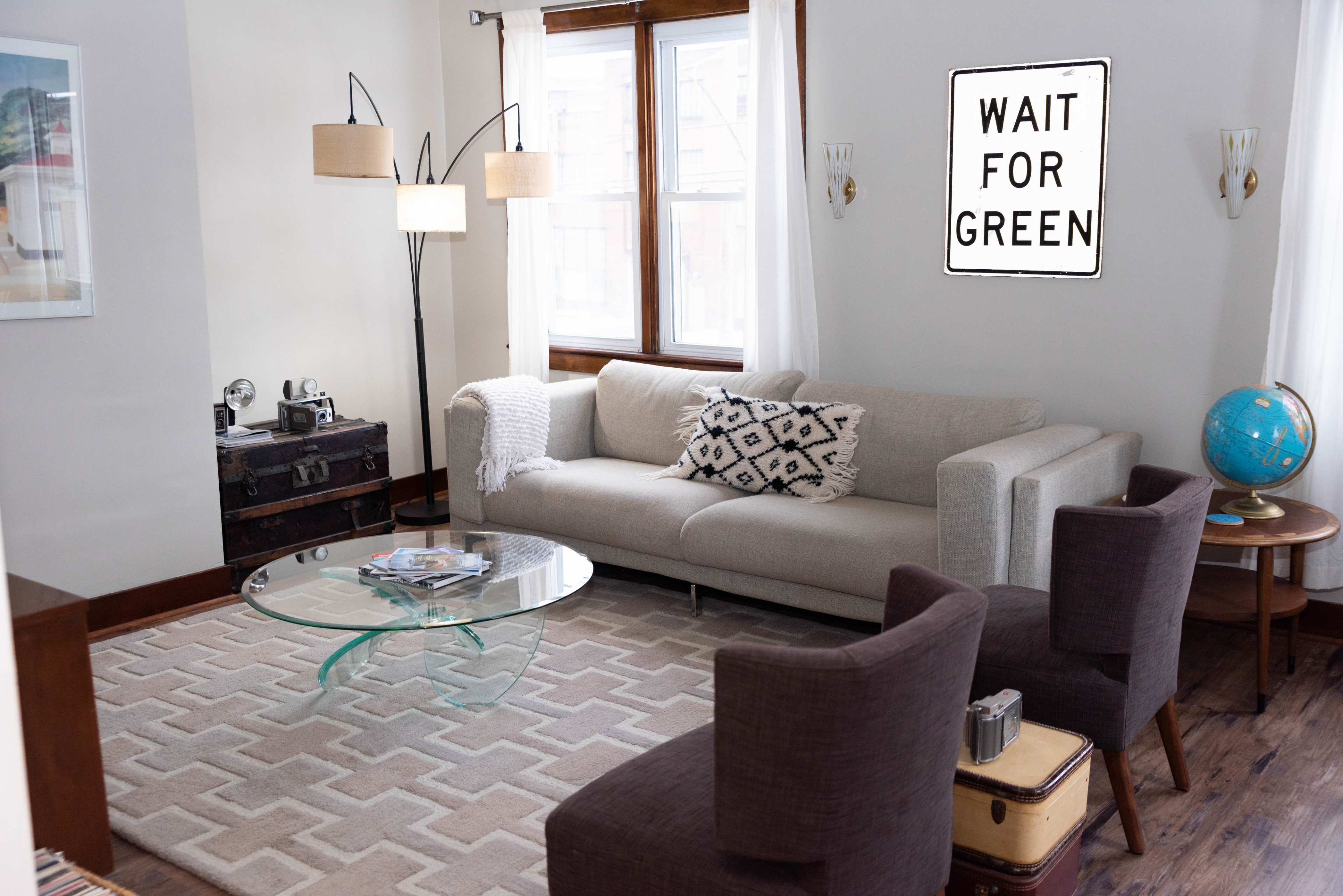 The living room features a light gray sofa, two dark chairs, a glass coffee table, and a decorative sign on the wall that reads "WAIT FOR GREEN."