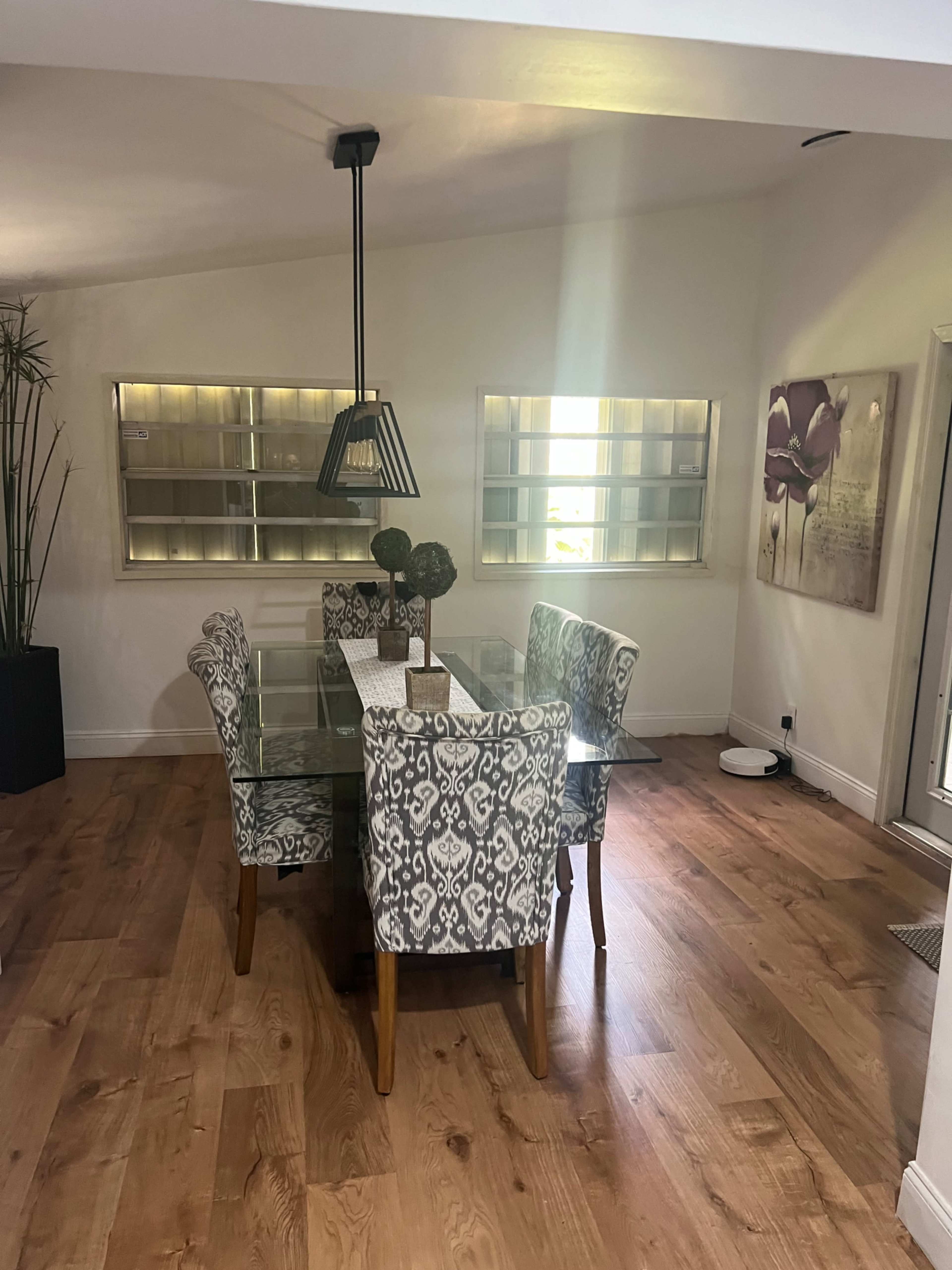 A dining area features a glass-top table surrounded by patterned chairs, illuminated by a pendant light, with decorative plants and wall art in the background.