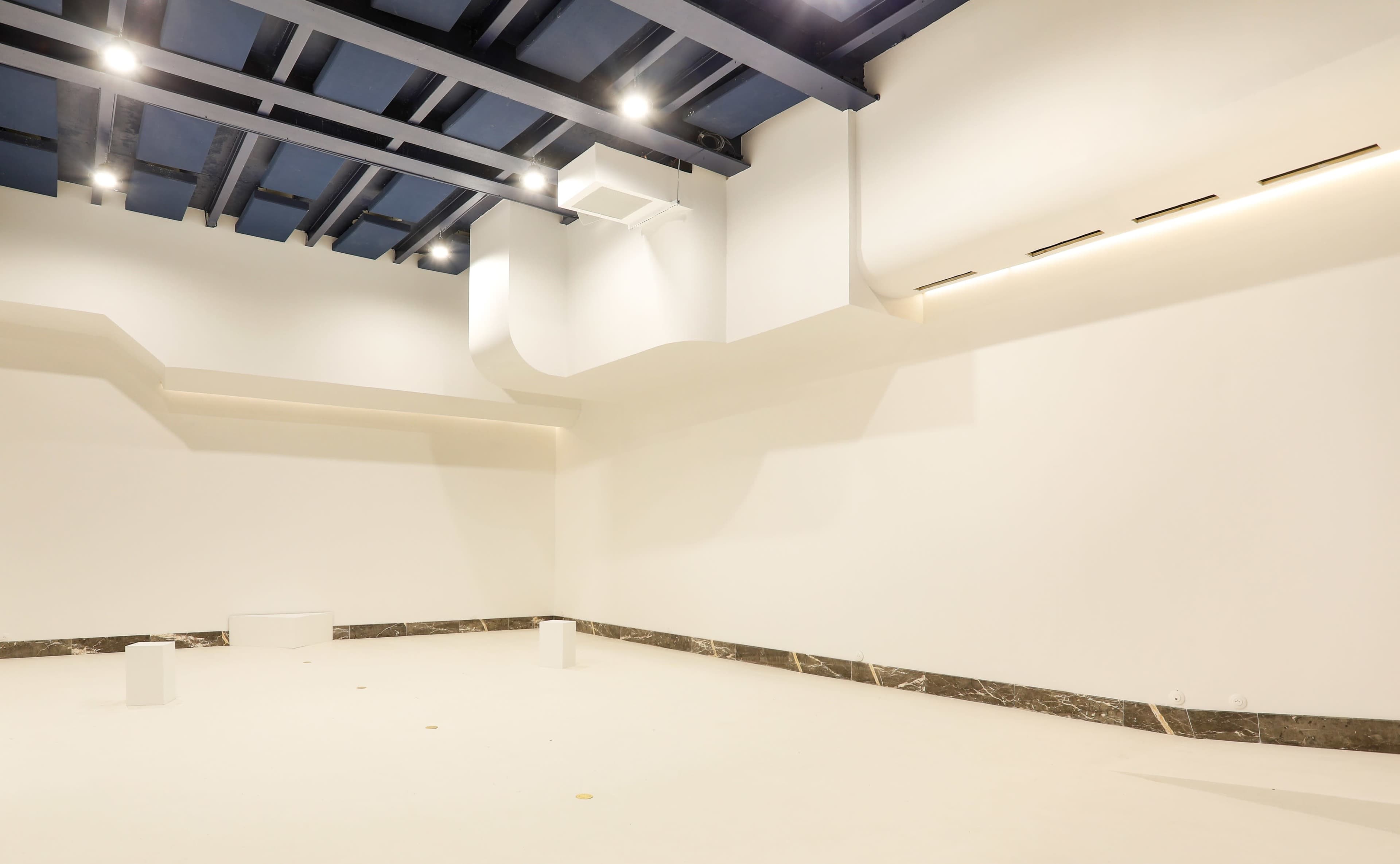 A minimalist interior space with white walls, a light-colored floor, and blue ceiling beams, featuring a few white cylindrical forms scattered throughout.