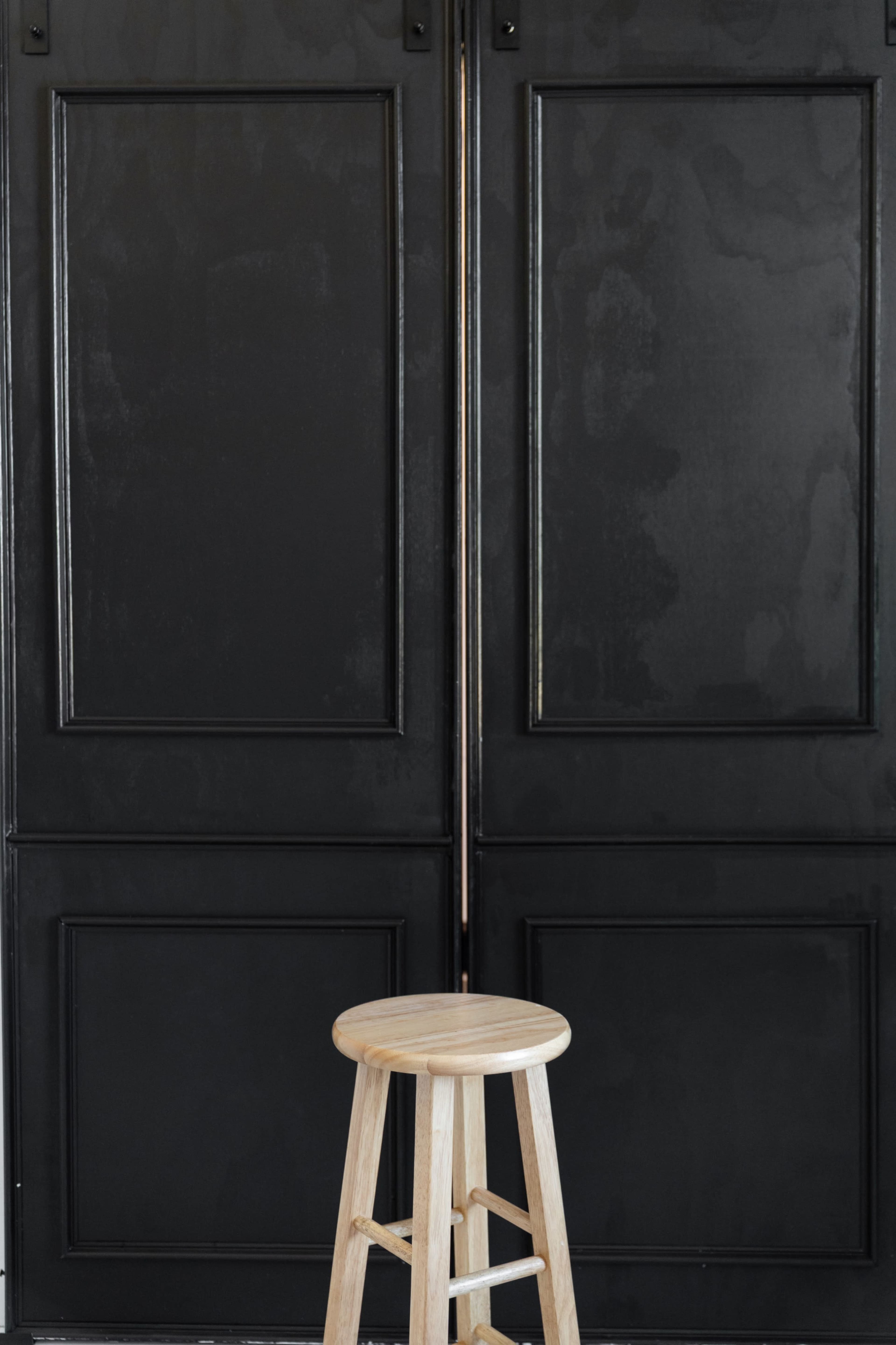 The image shows a wooden stool positioned in front of a pair of large black sliding doors with a flat panel design.