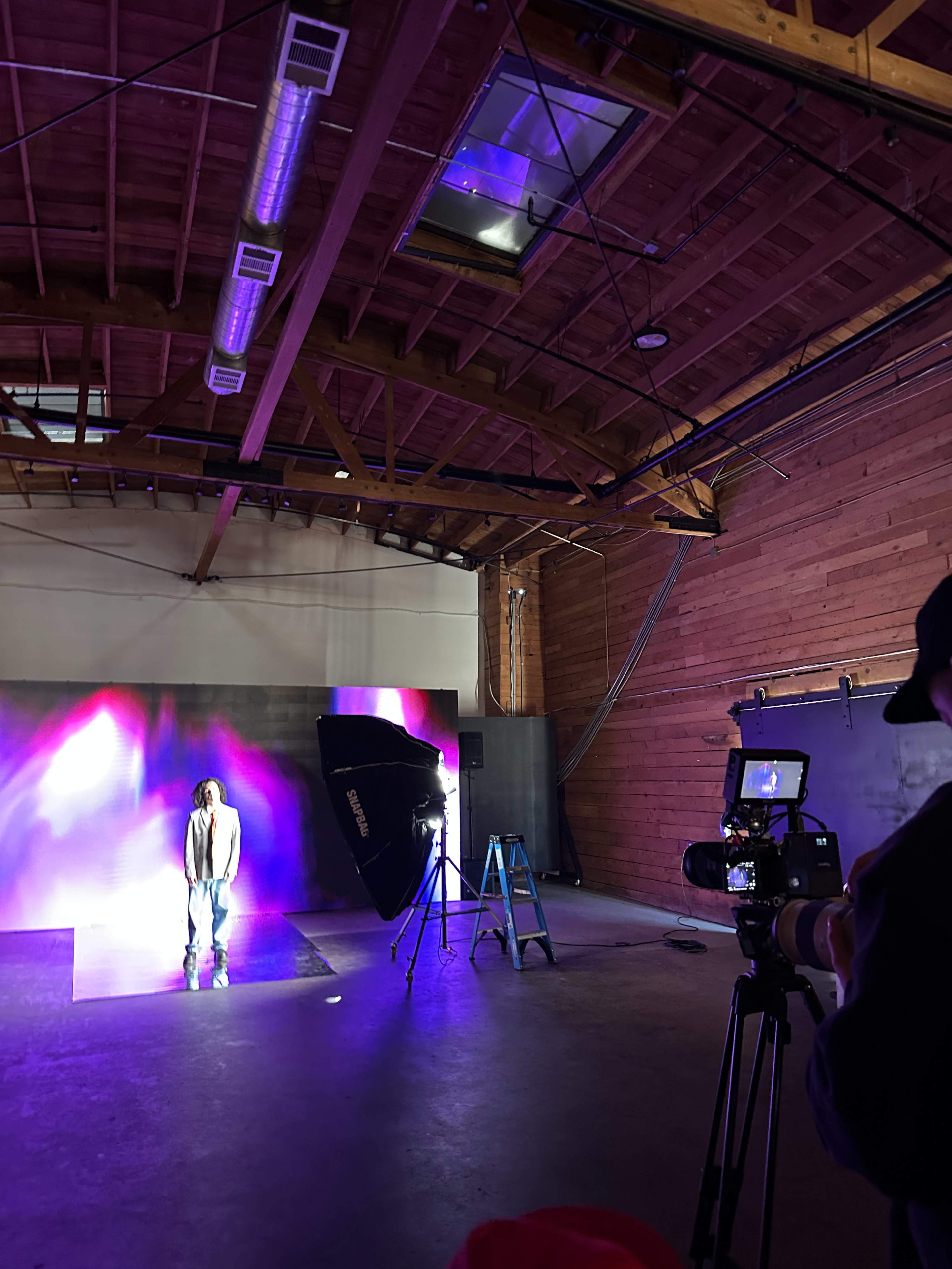 A person stands on a reflective surface in an industrial studio, while a camera operator and lighting equipment are positioned nearby.