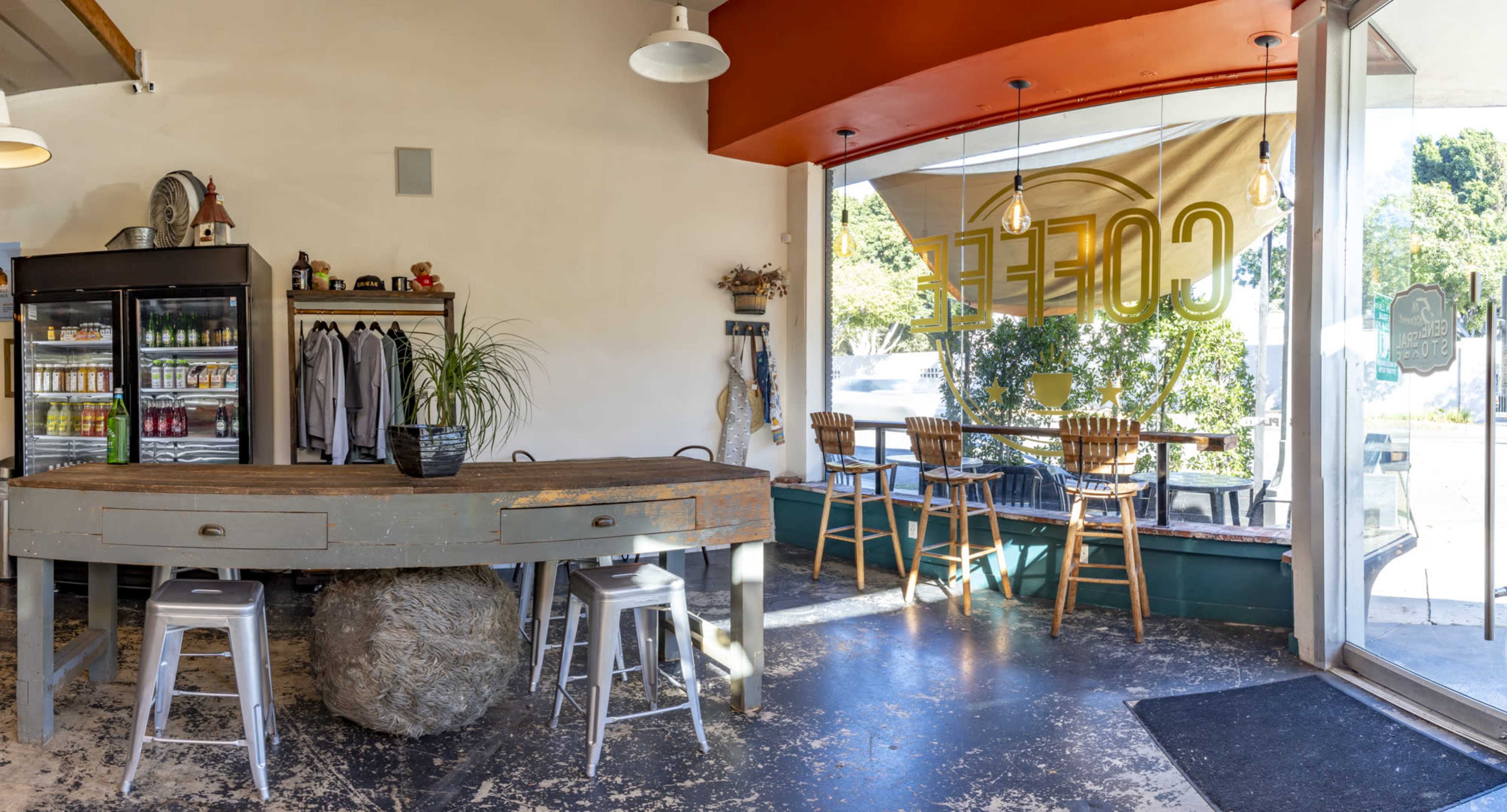 A coffee shop interior features a large round wooden table surrounded by metal stools, a glass display case filled with drinks, and tables near the window overlooking the outside.