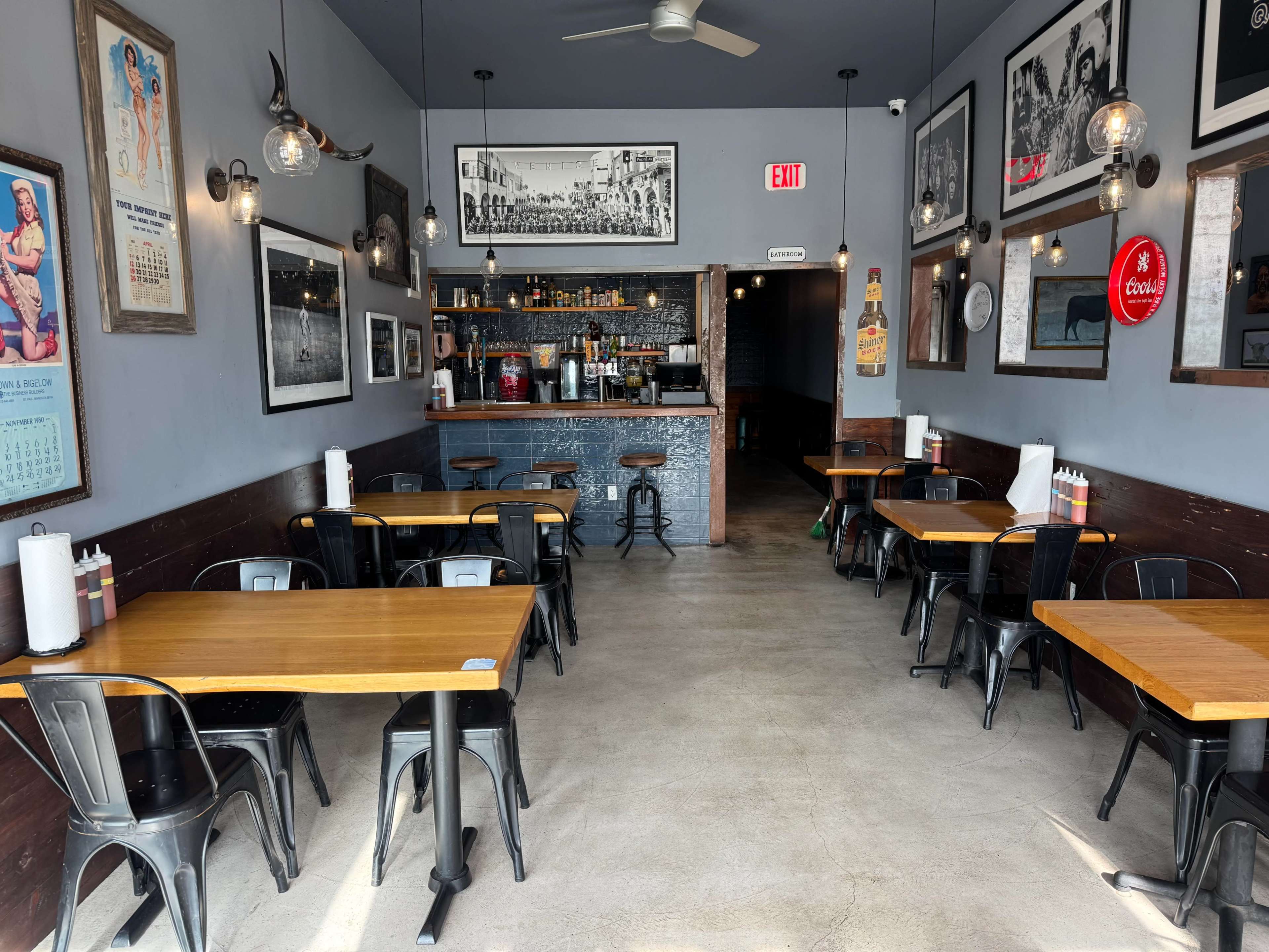 The image shows a restaurant interior with wooden tables and black metal chairs arranged around the space, featuring a bar area in the back and framed photographs on the walls.