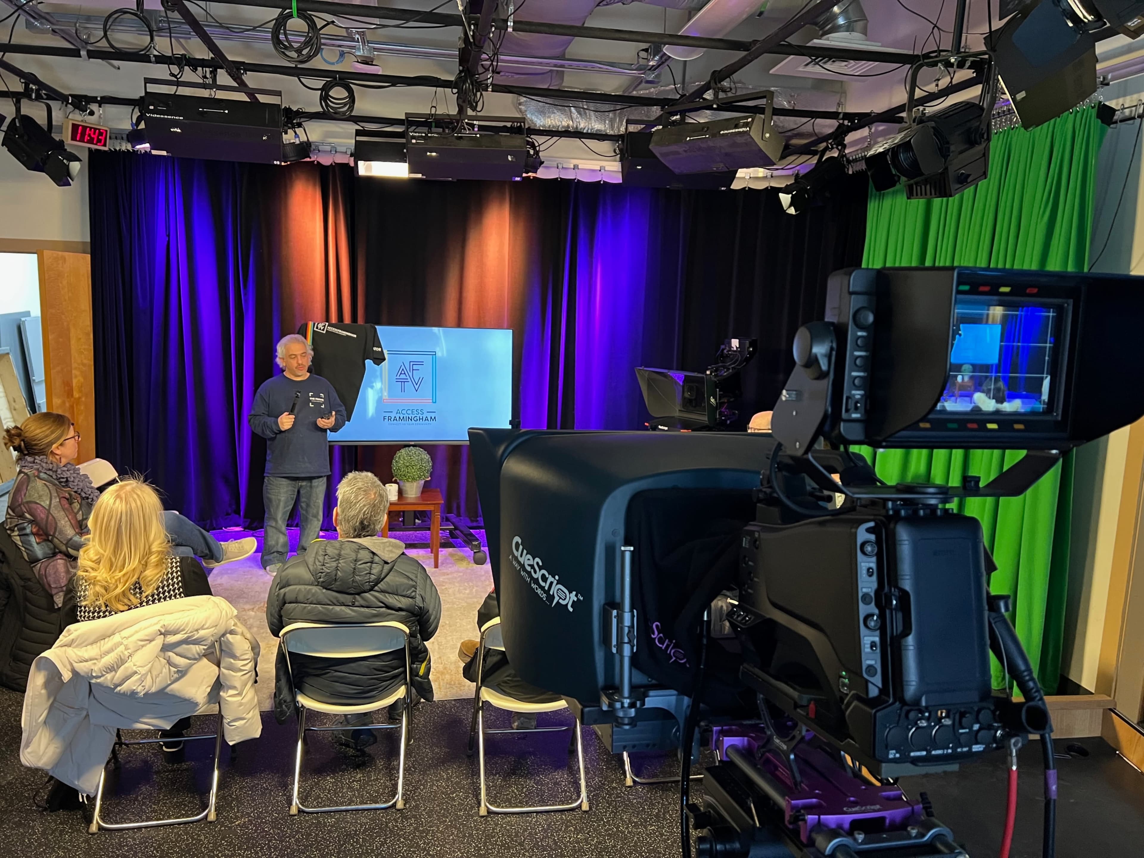A speaker presents in a studio setting with an audience seated in front of him, surrounded by video equipment and colorful lighting.