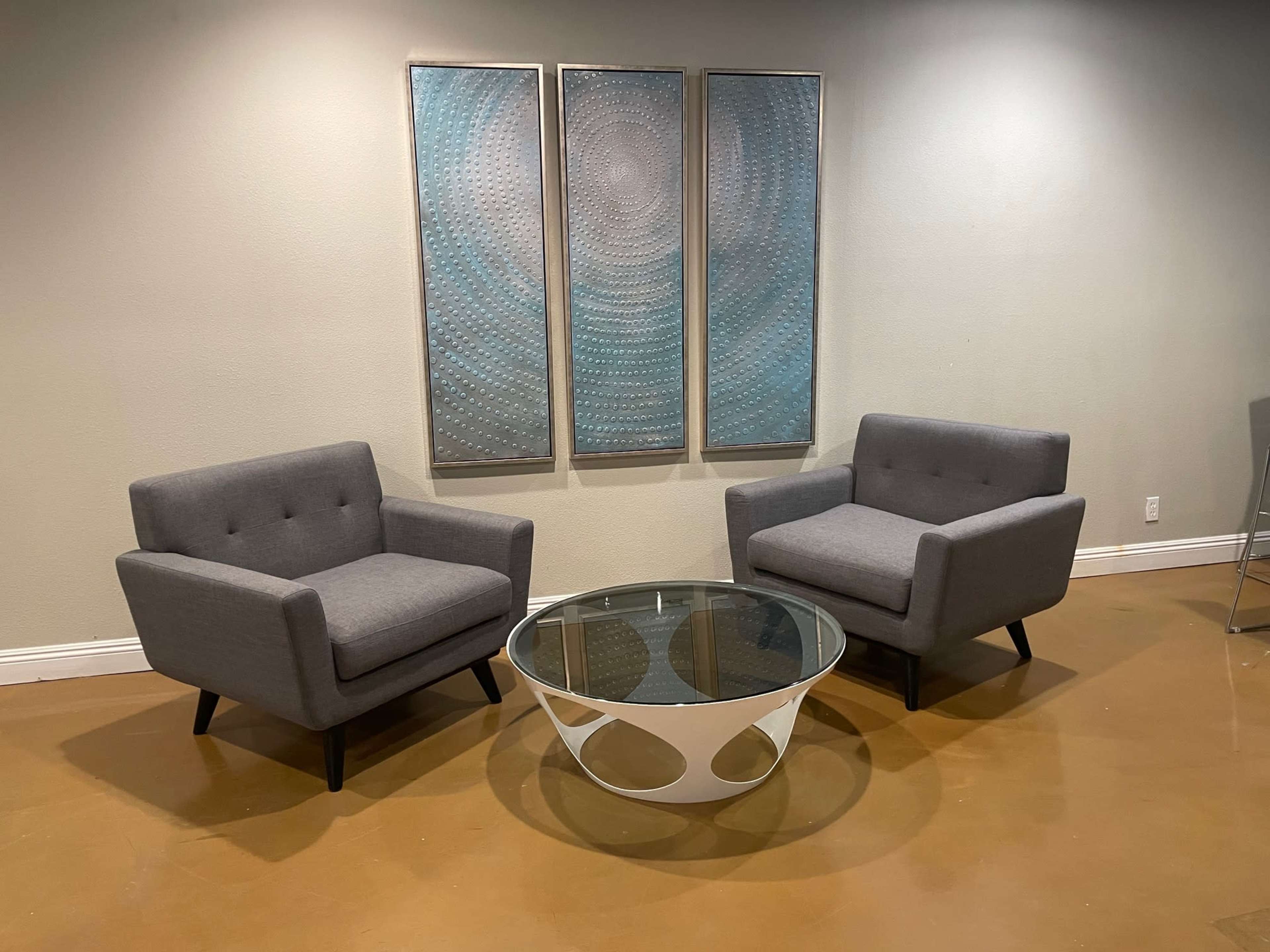 Two gray armchairs facing each other around a round glass coffee table, positioned in a modern room with light-colored walls and abstract wall art.