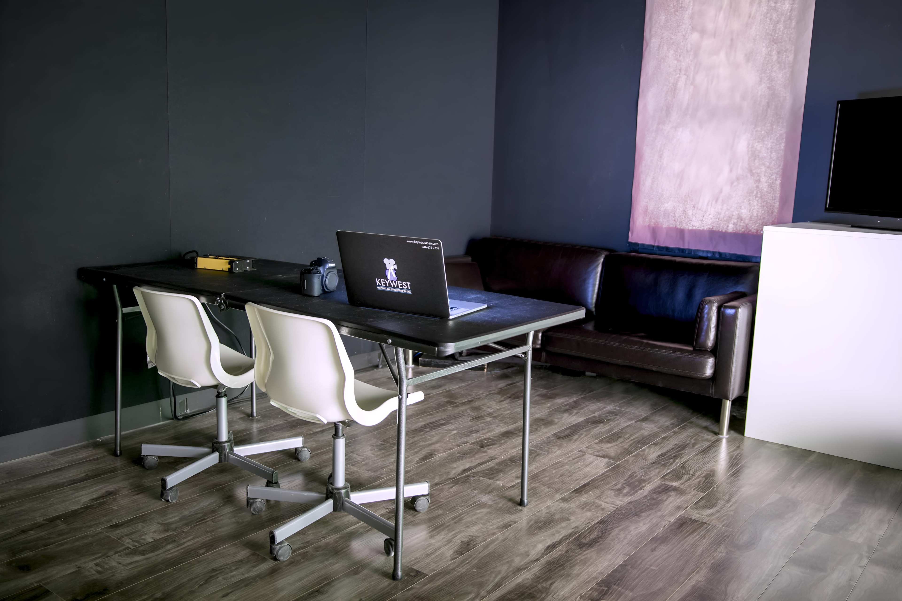 A modern office space with a black table, three white chairs, a laptop on the table, and a brown leather couch against the wall.
