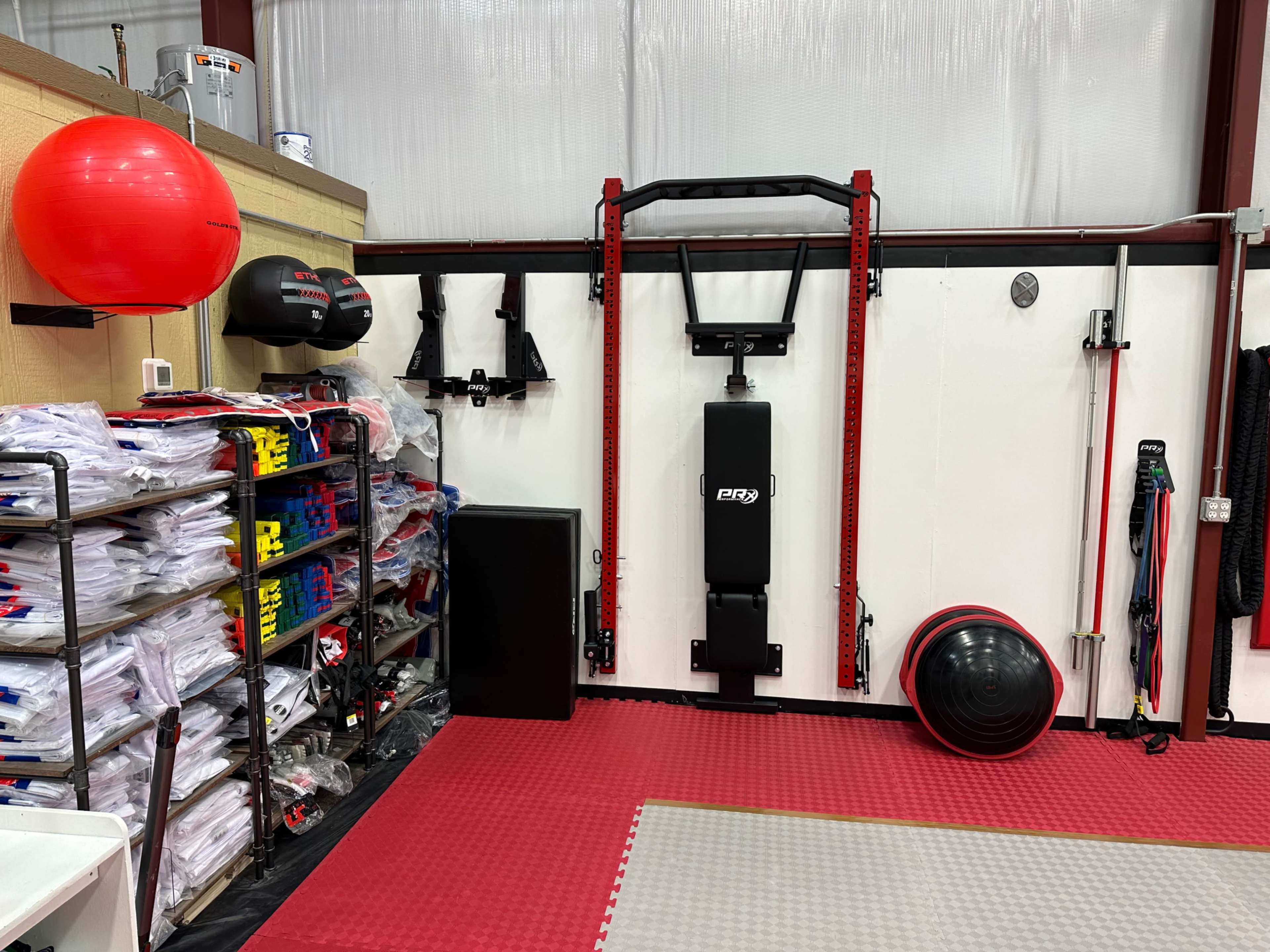 A fitness area with various exercise equipment, including a strength training station, workout mats, and storage shelves filled with white training uniforms.