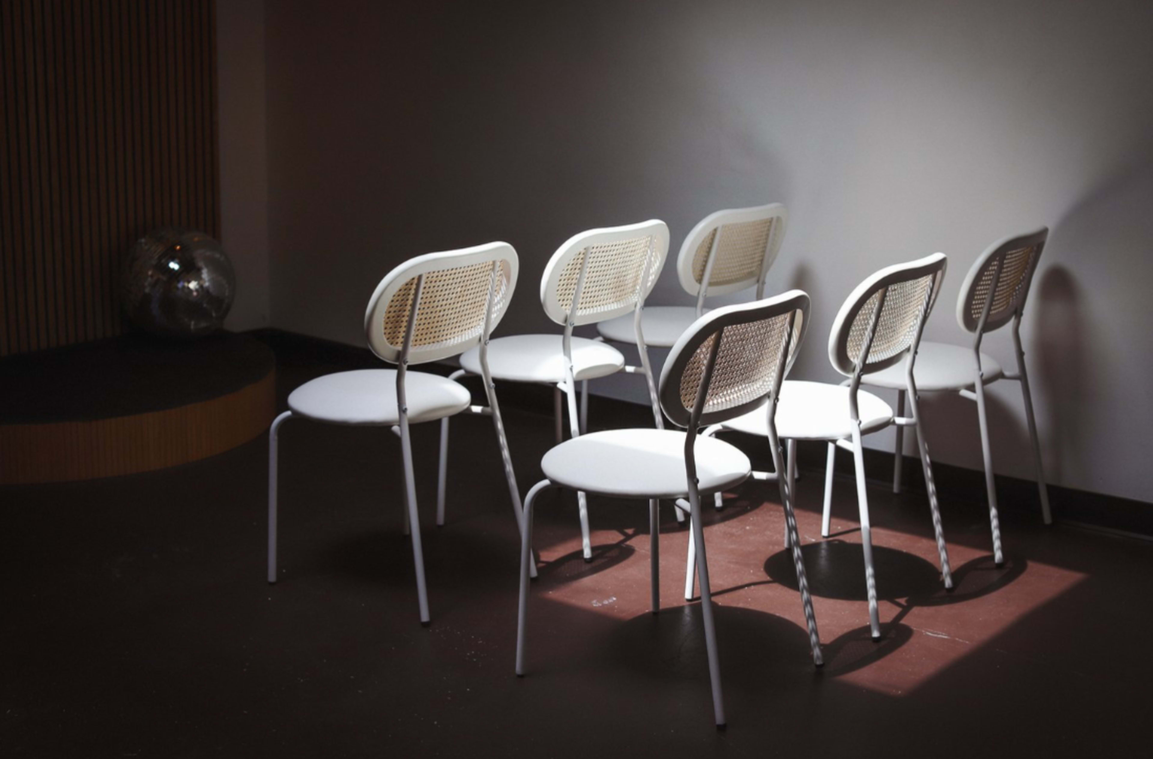 A row of white chairs with woven backs is arranged in a dimly lit room.