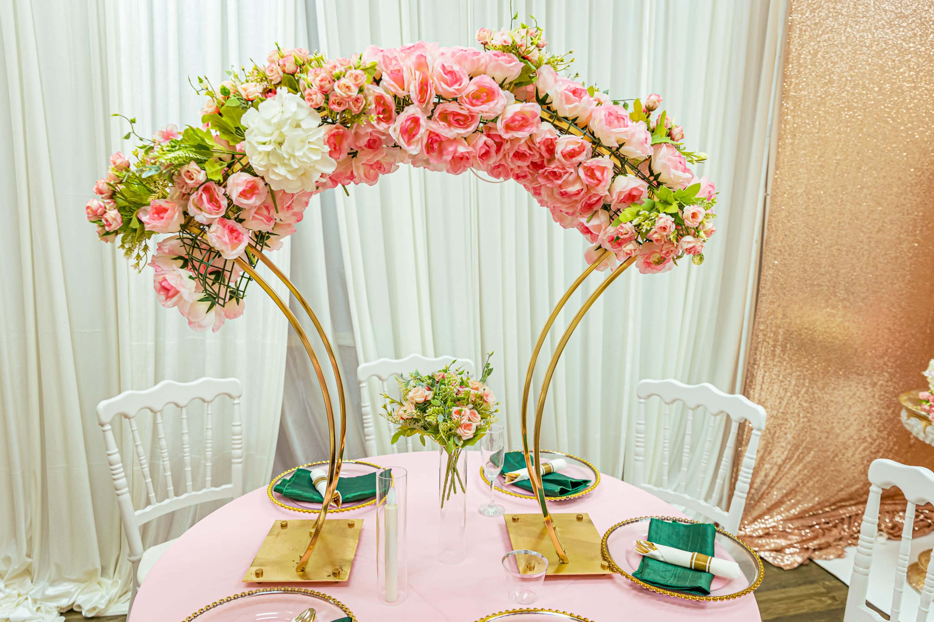 A table is set for an event with a floral arch made of pink roses, placed over a pink tablecloth and complemented by decorations and table settings.