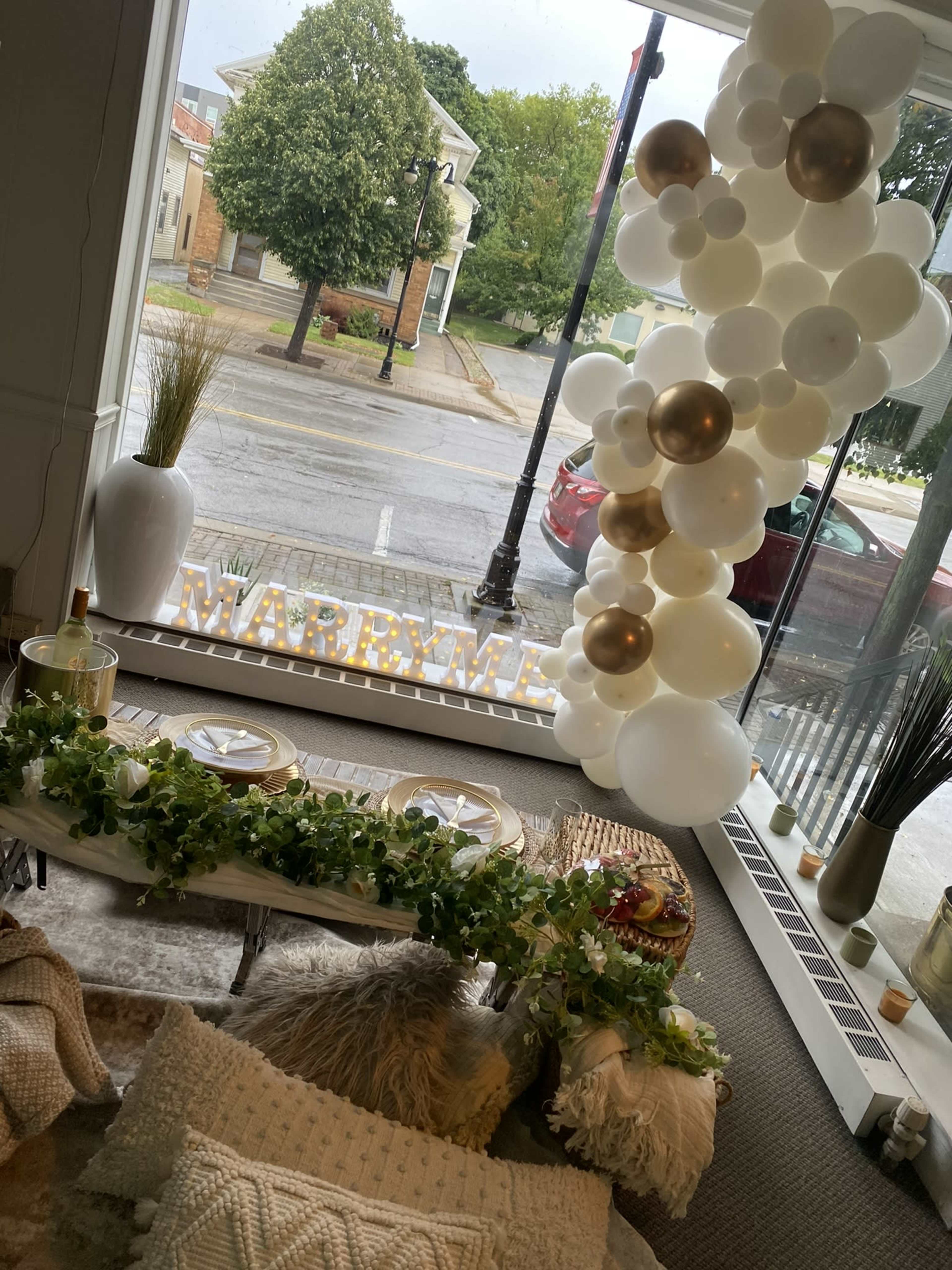 The interior showcases a decorative setup for a celebration, featuring a table with greenery, white and gold balloons, and illuminated letters spelling "MARRY ME" against a window view.