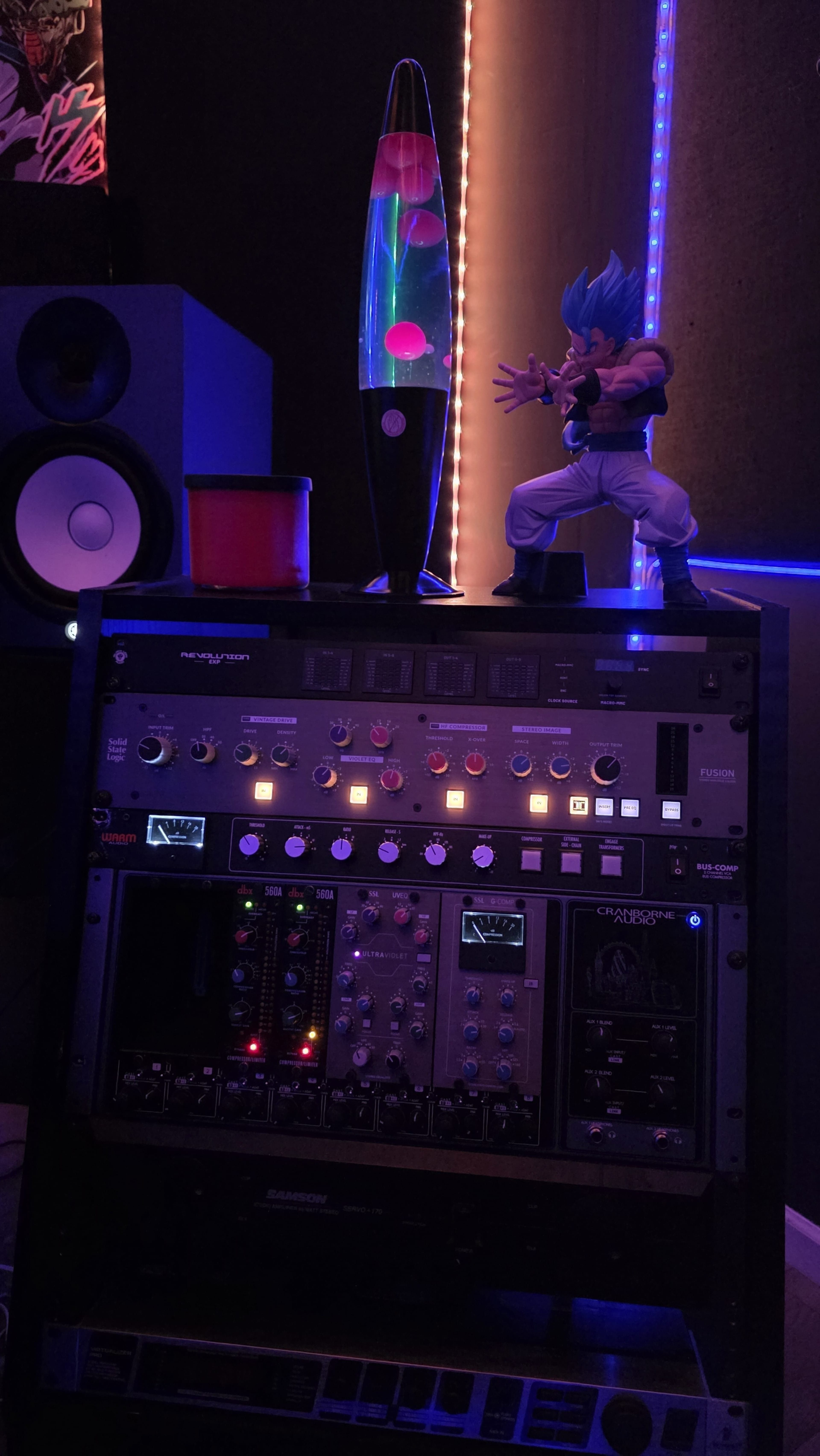 The image features a recording equipment rack with various audio controls, a lava lamp, and a figurine of a character in a dynamic pose, set against a backdrop of colored lights.