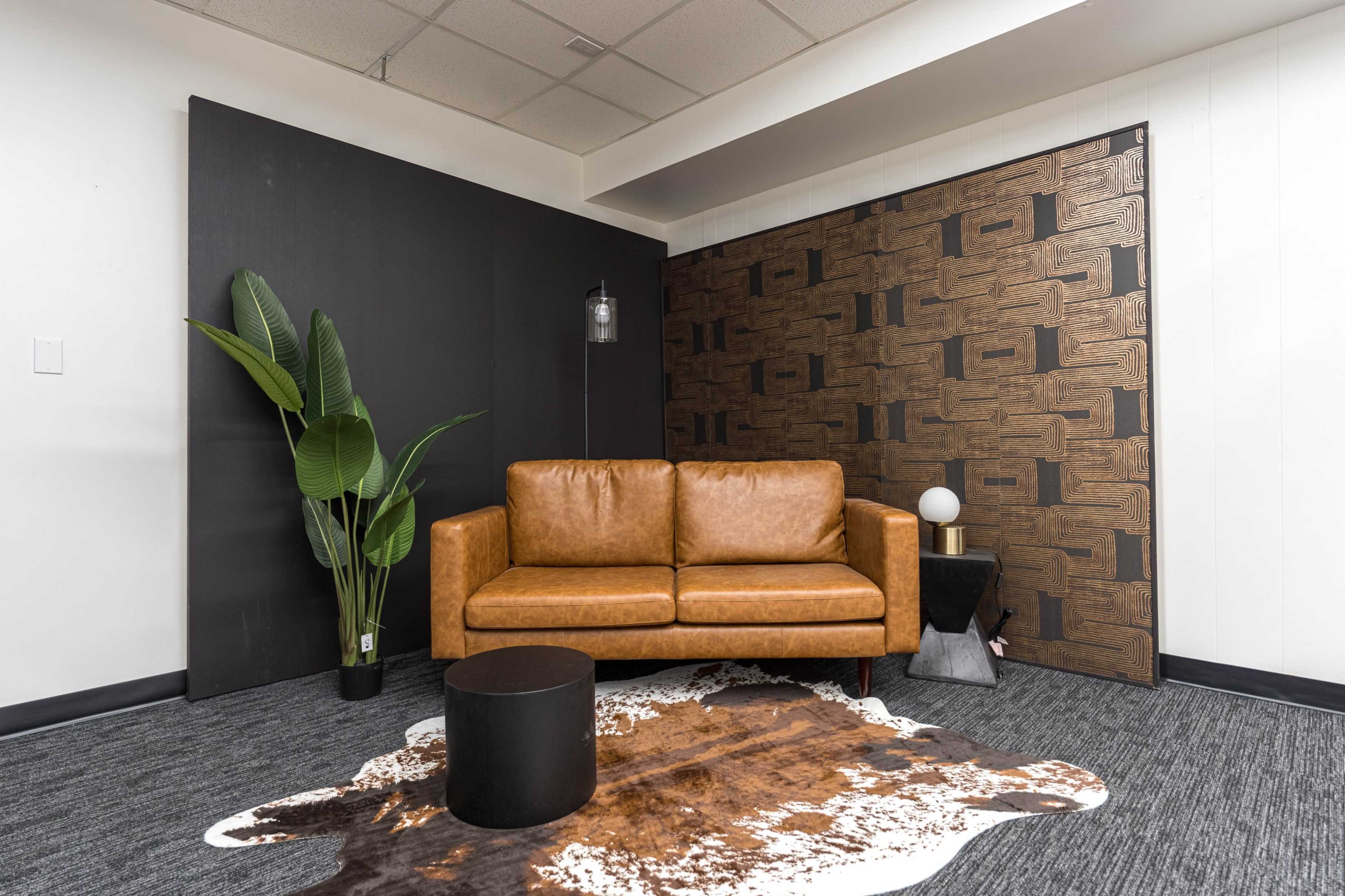 A brown leather sofa is placed in a minimalist room with a dark wall and a textured decorative wall, accompanied by a small black table and a large plant.