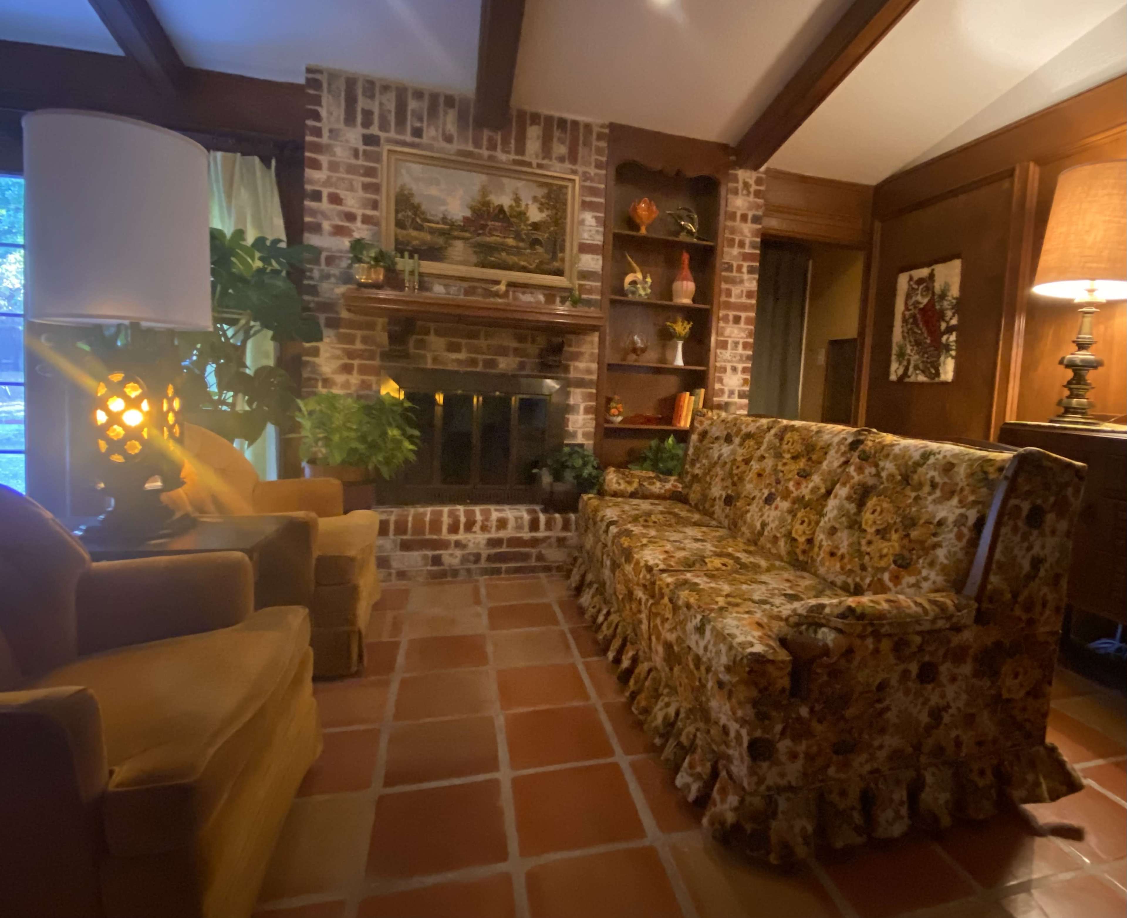 The image shows a cozy living room featuring a floral-patterned sofa, two armchairs, a brick fireplace, and wooden shelves with decorative items.