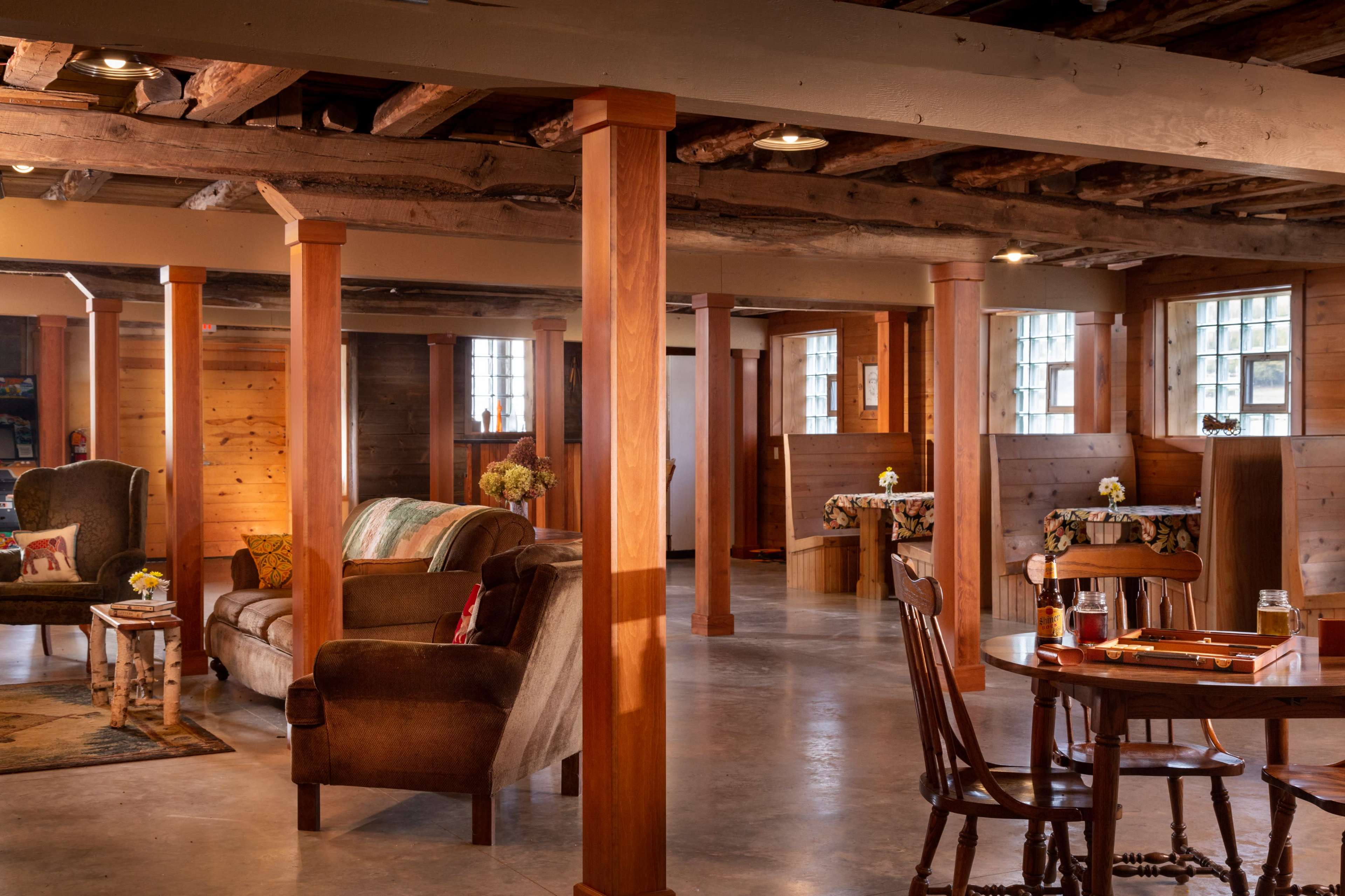 The image shows a spacious, wooden-paneled room with several seating areas, light wooden columns, and tables adorned with tablecloths.