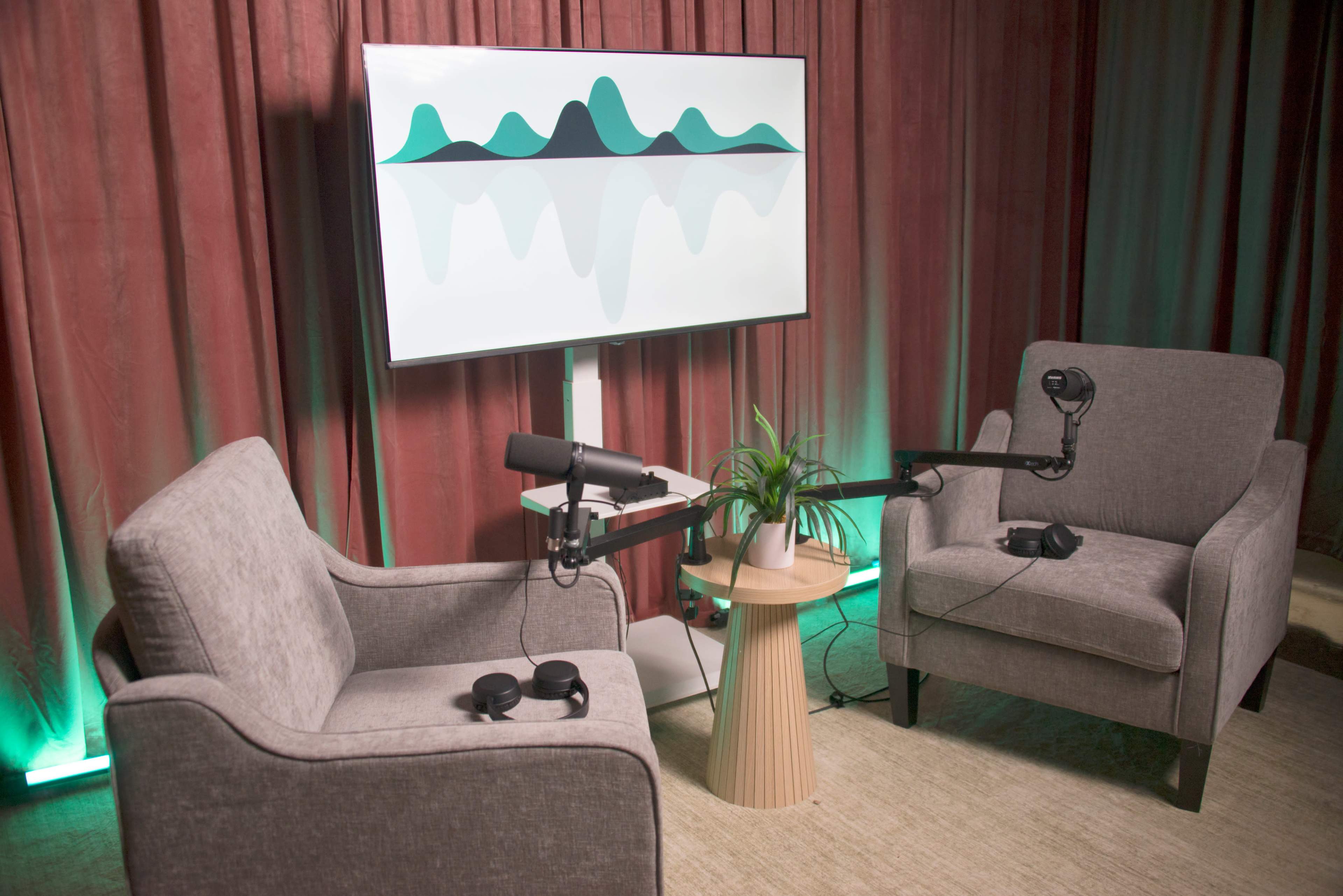 The image shows a minimalist podcasting setup with two gray armchairs, a small table, a microphone mounted on a stand, headphones on each chair, and a large screen displaying abstract graphic mountains in the background.