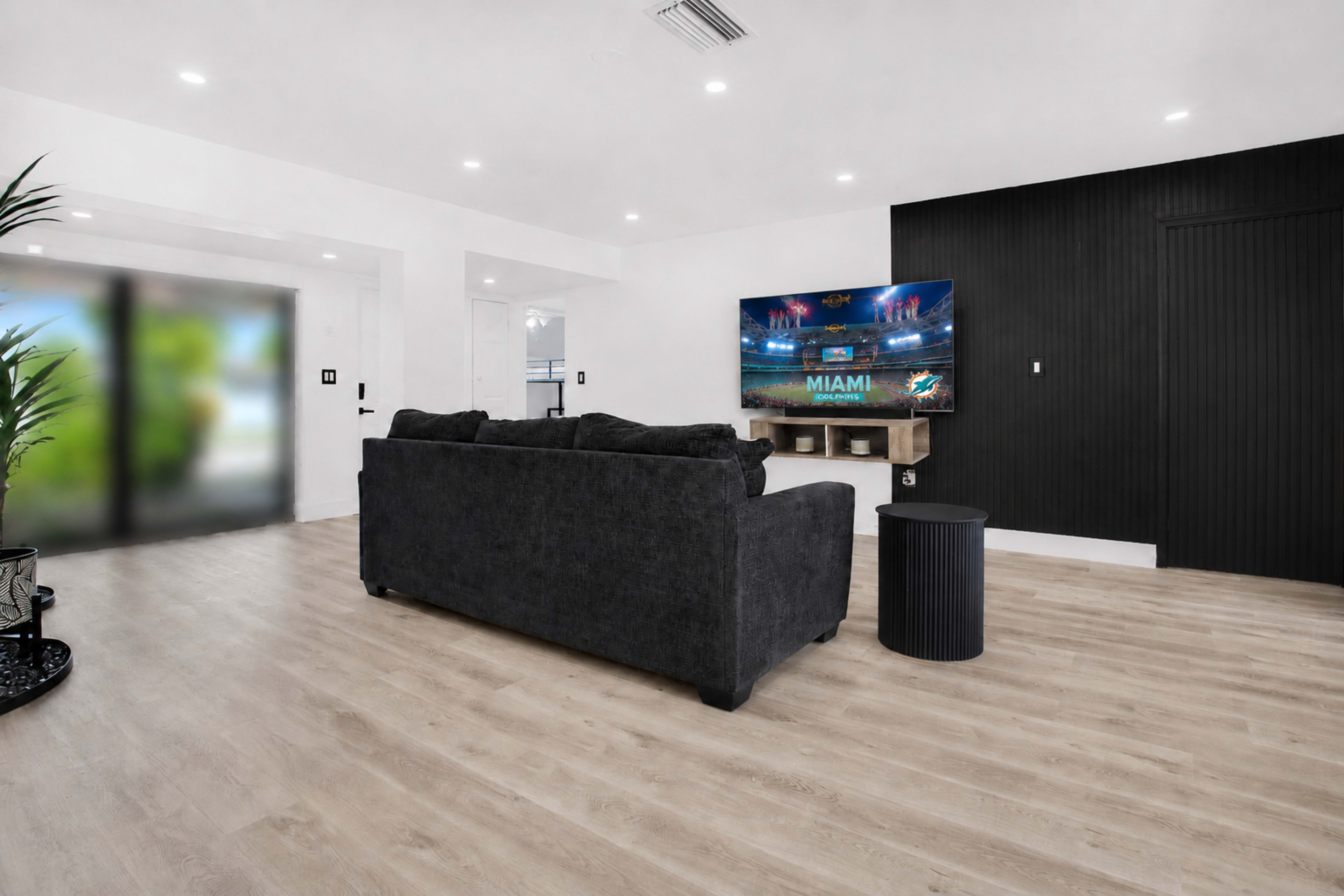 A dark couch faces a mounted television in a modern living room with light wood flooring and minimalistic decor.