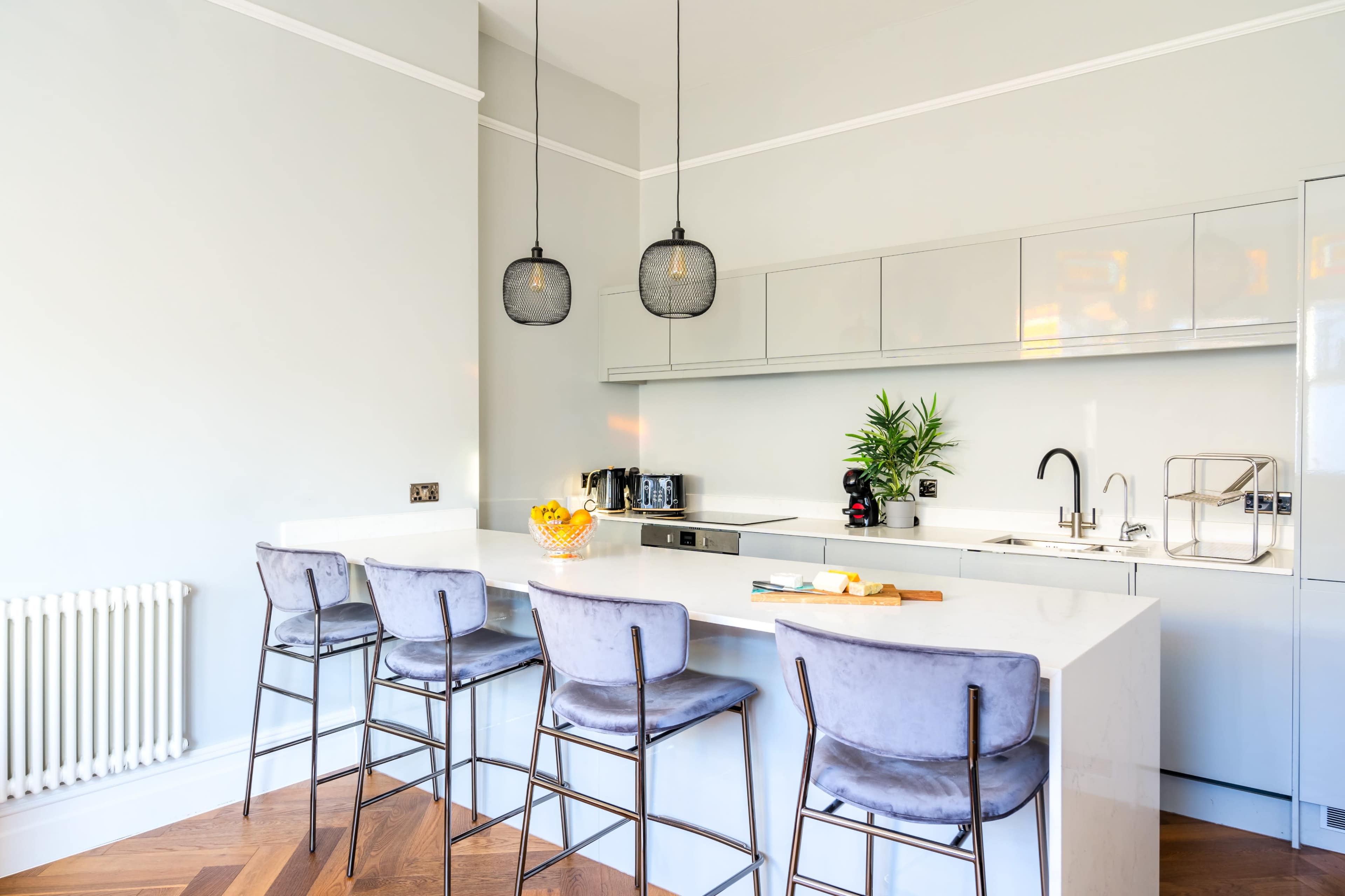 A modern kitchen features a white countertop with four high-backed stools, pendant lights overhead, and minimalist cabinetry along the wall.