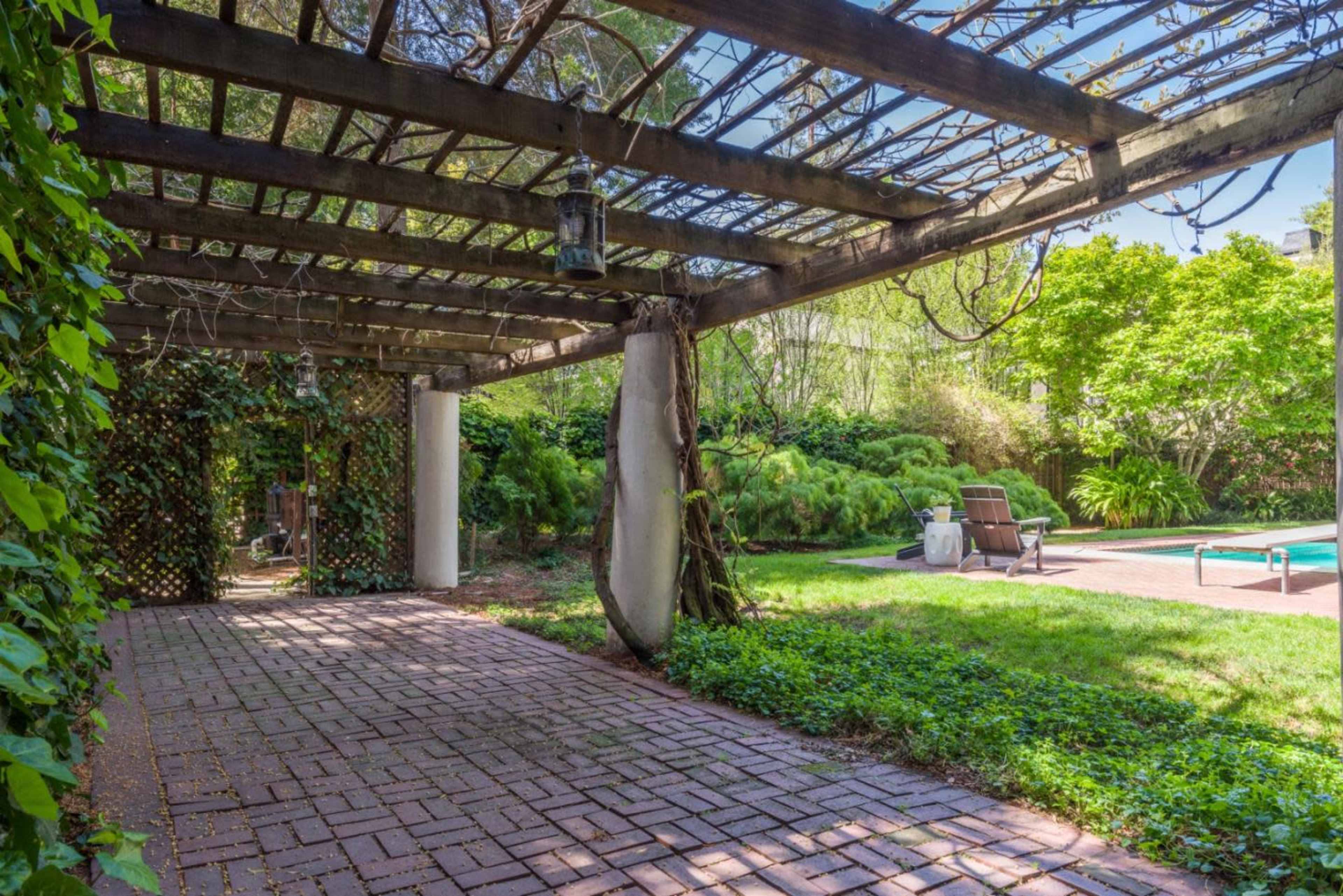 A brick patio with a wooden pergola covered in greenery overlooks a pool area surrounded by lush plants.