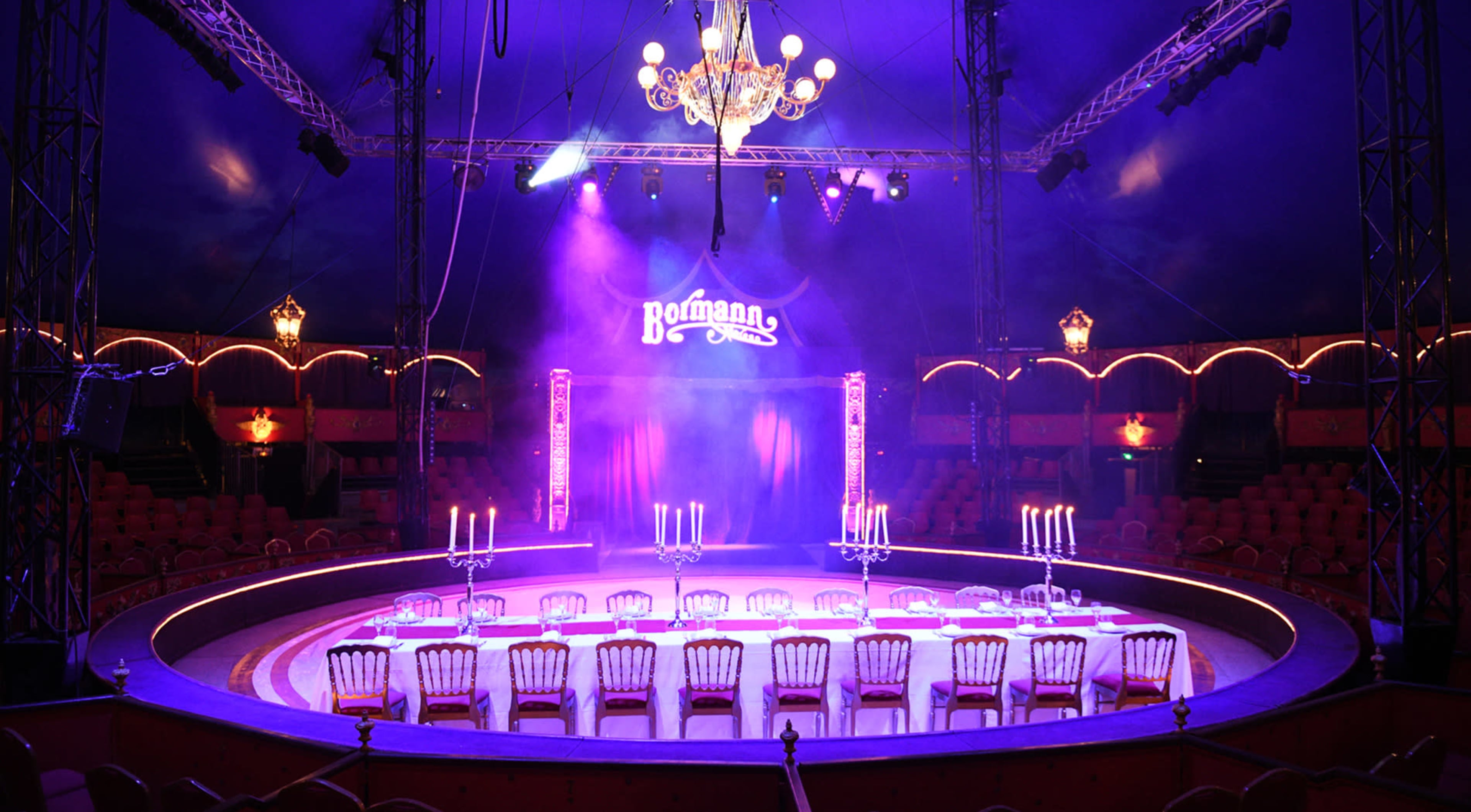 A brightly lit circular stage set up with a long table and chairs, surrounded by empty seats in a circus tent.