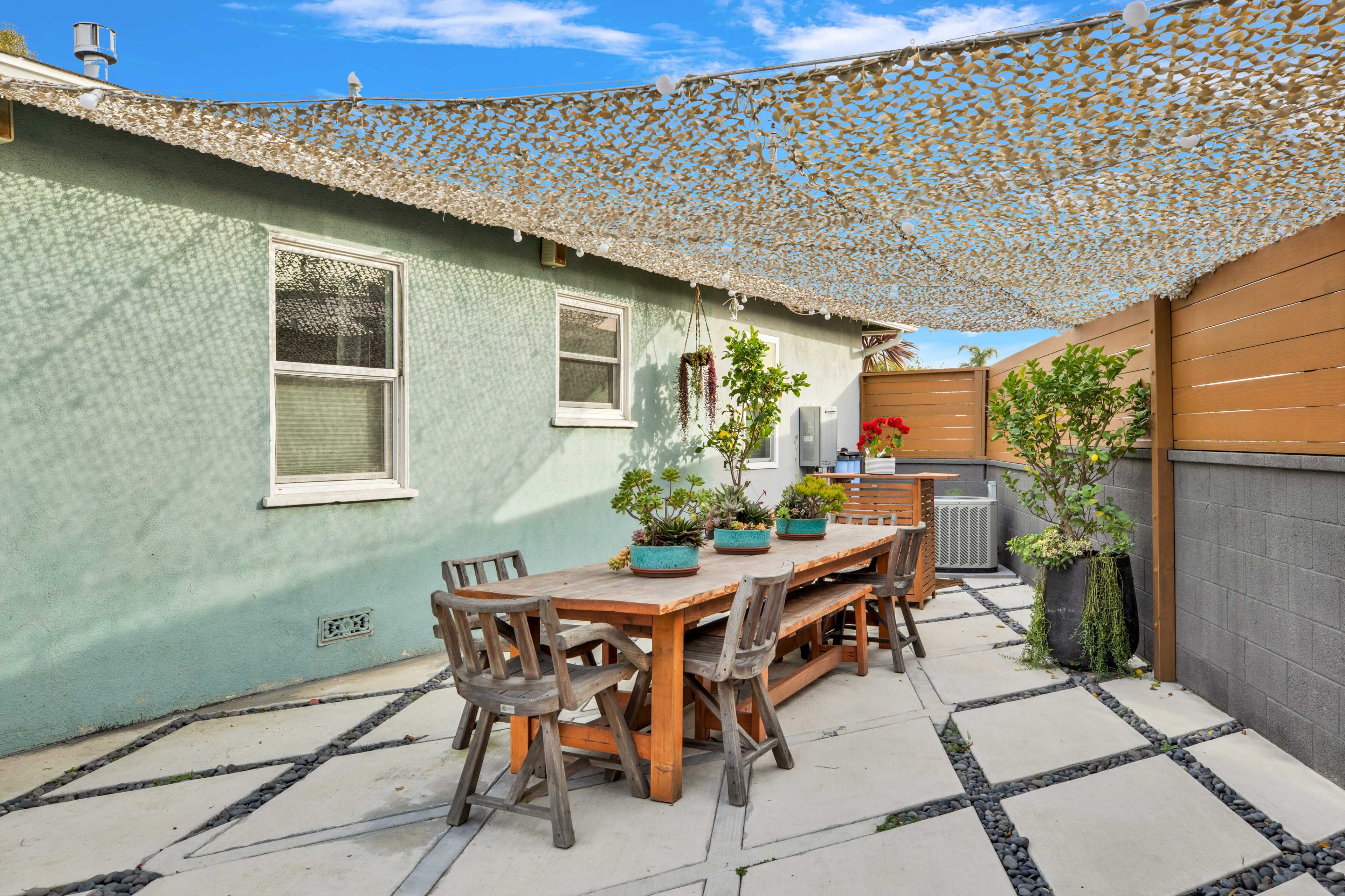A wooden table surrounded by chairs sits under a shaded canopy in a patio area with potted plants.