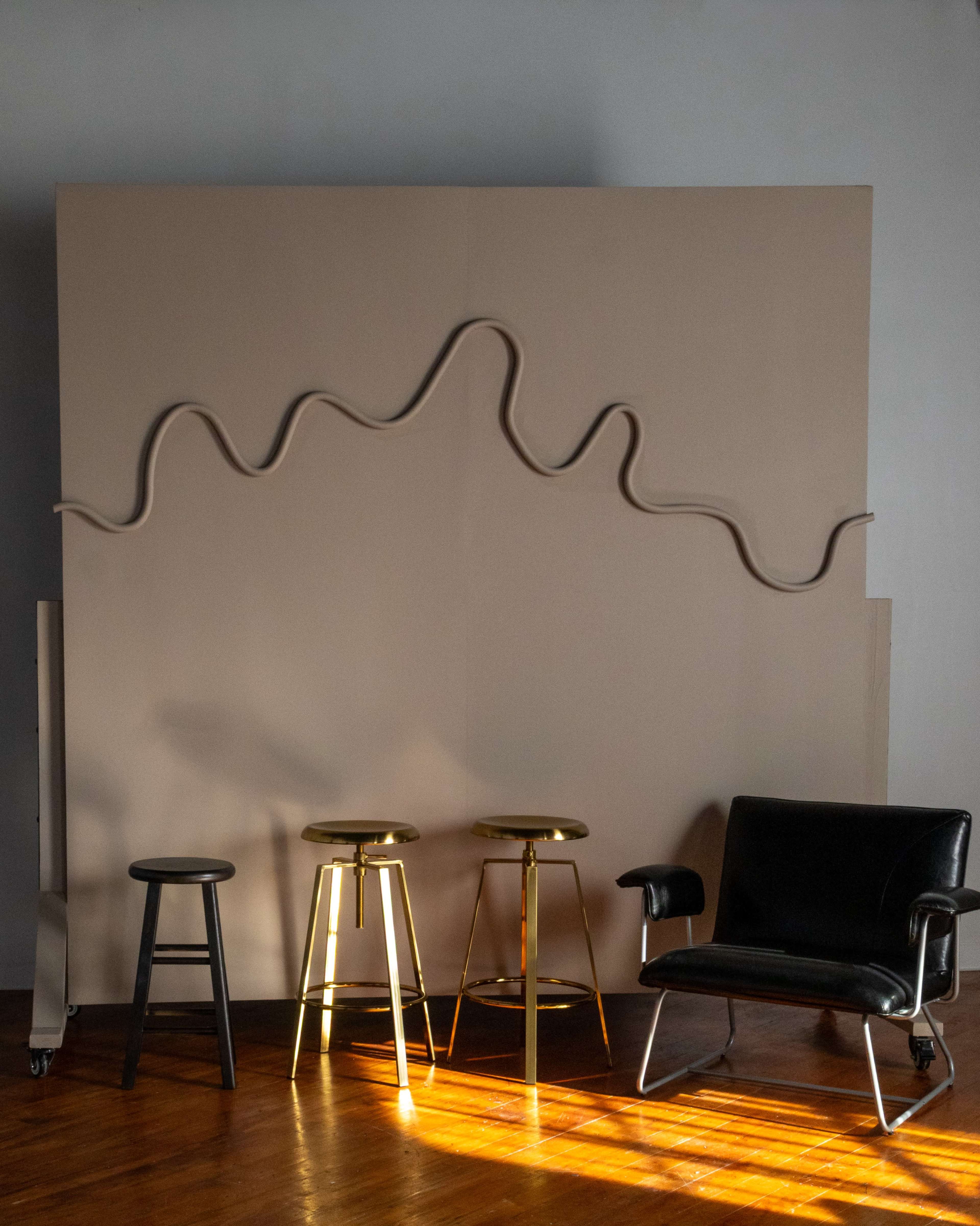 The image shows a minimalistic interior with three metal bar stools and a black chair against a large beige wall featuring a wavy sculpture.