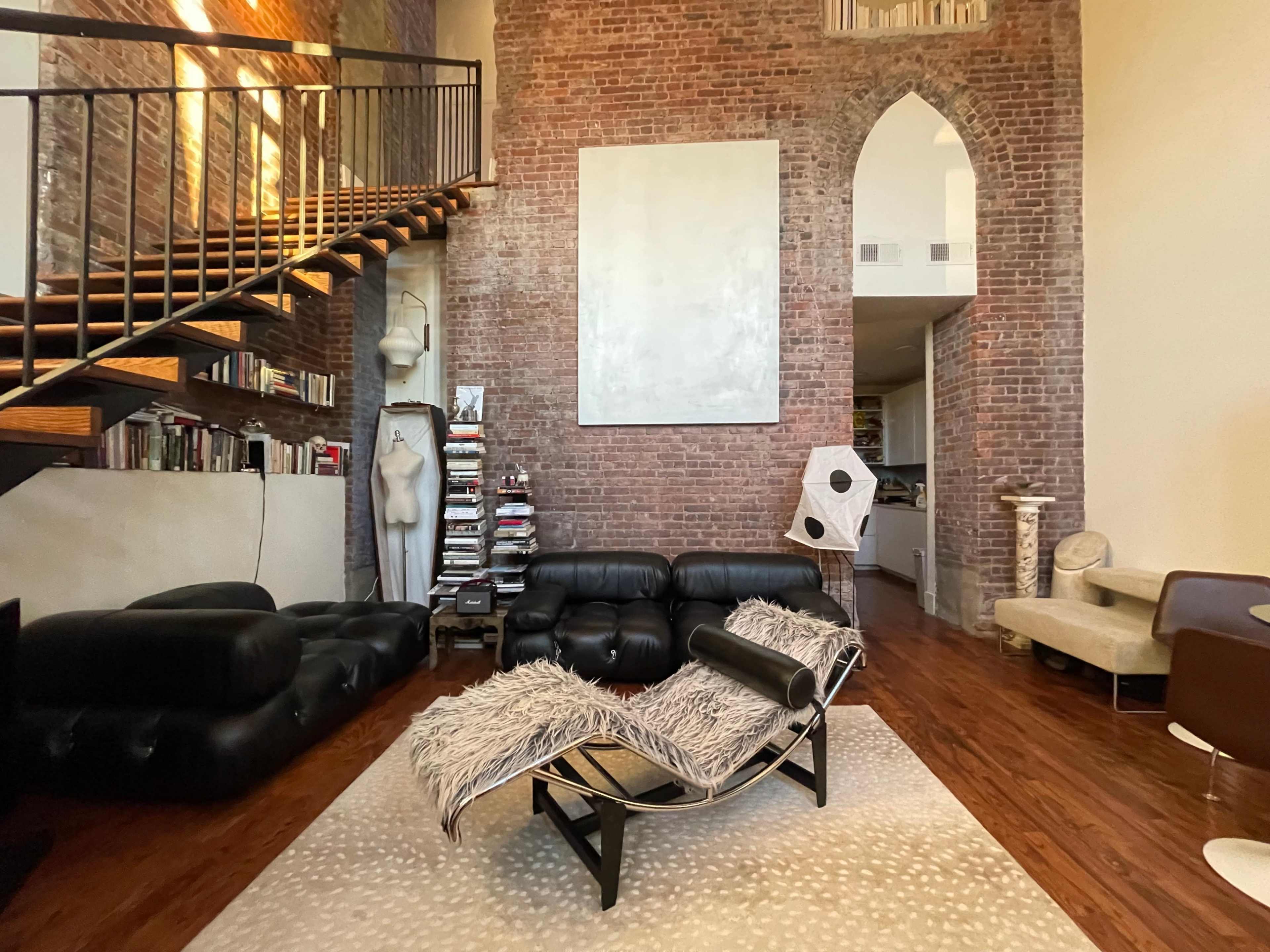 The room features a modern lounge area with a black leather sofa, a unique lounge chair, and a staircase leading to a loft, all set against exposed brick walls.