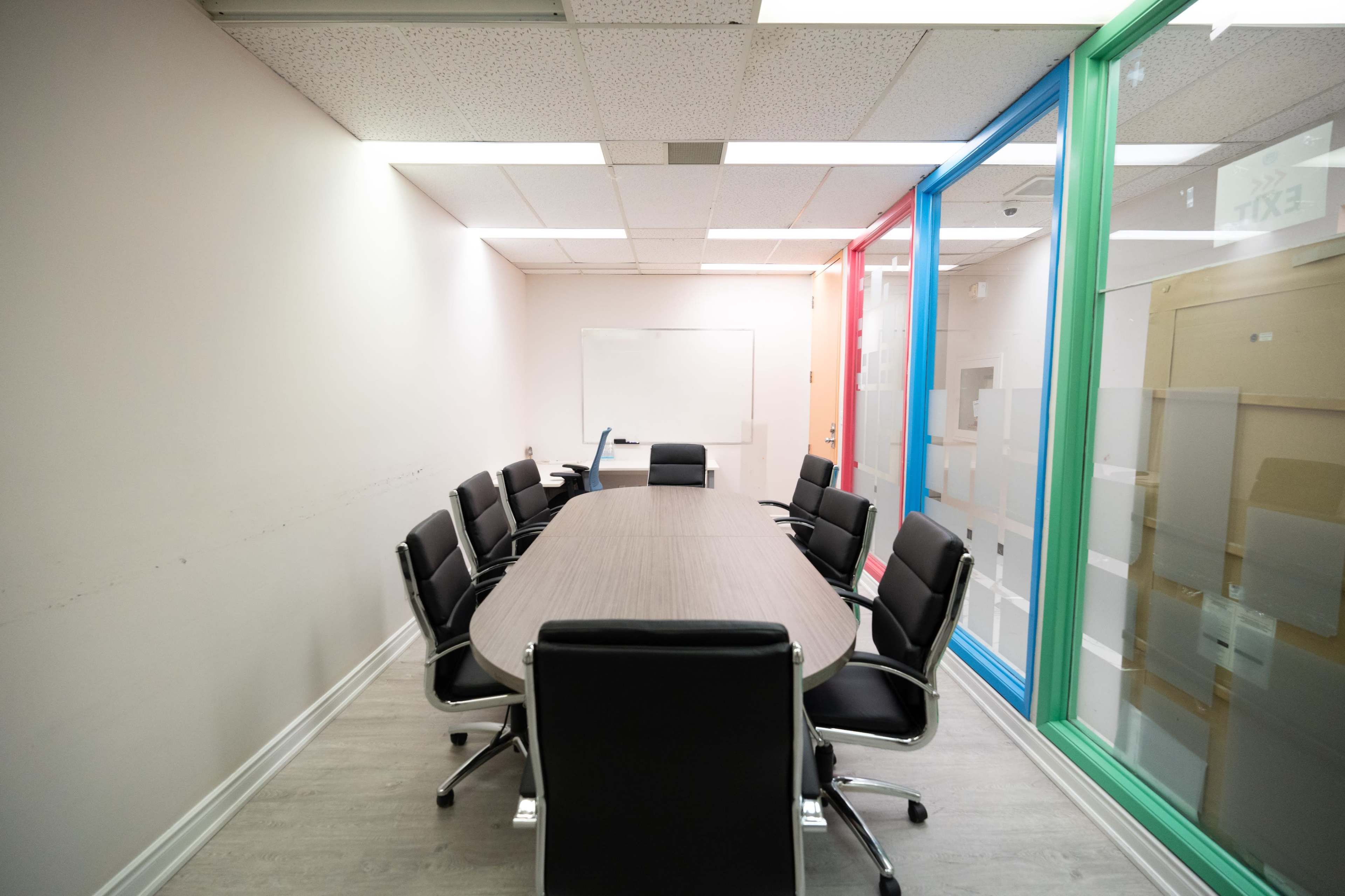 A long, oval conference table is surrounded by black chairs in a minimally decorated meeting room with colorful glass walls.
