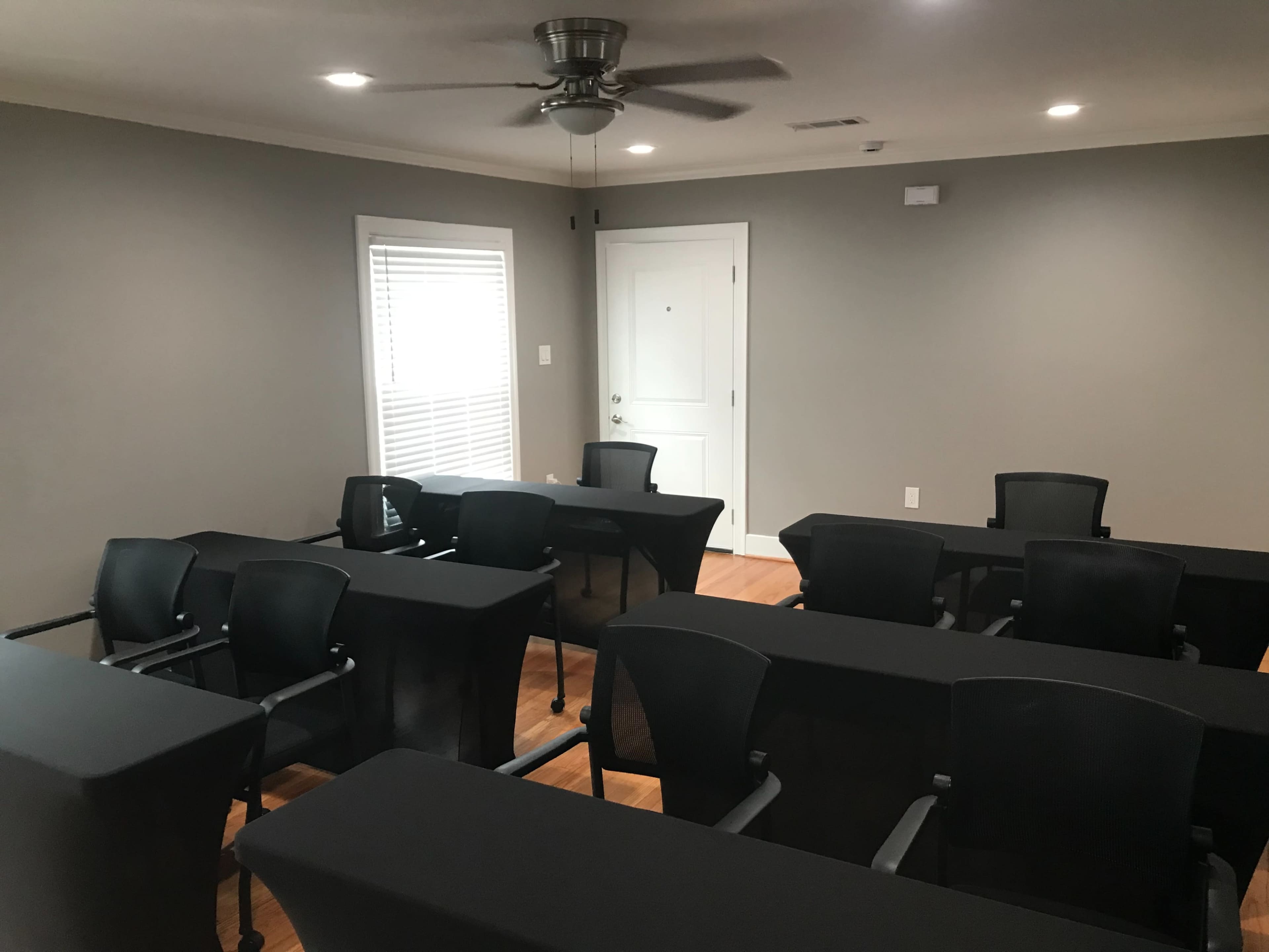 The image shows a conference room with several black tables and chairs arranged in a layout suitable for meetings, with a door and windows visible.