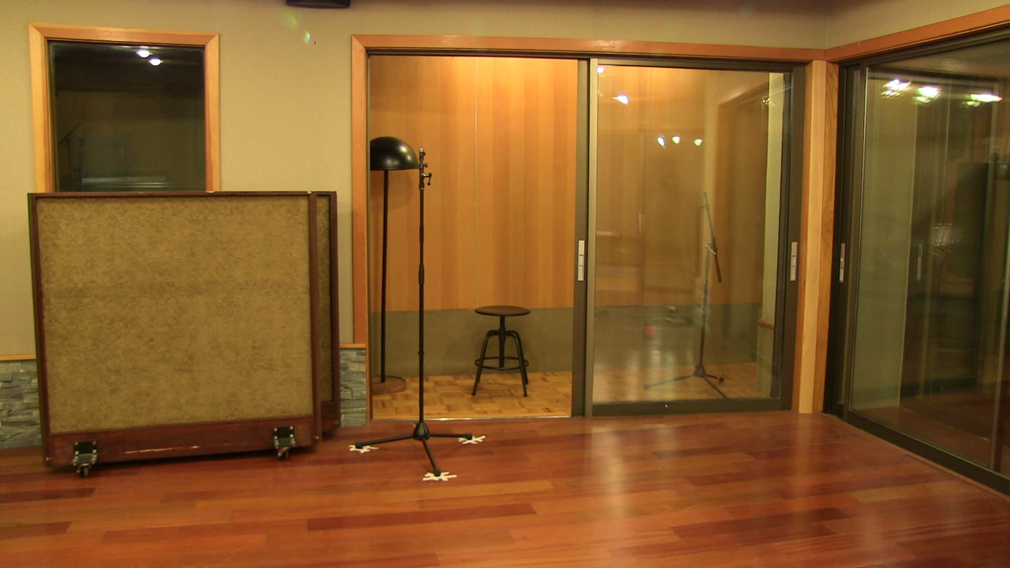 A sound studio with a glass-walled recording booth, a stool inside, and a microphone stand outside, set on polished wooden flooring.