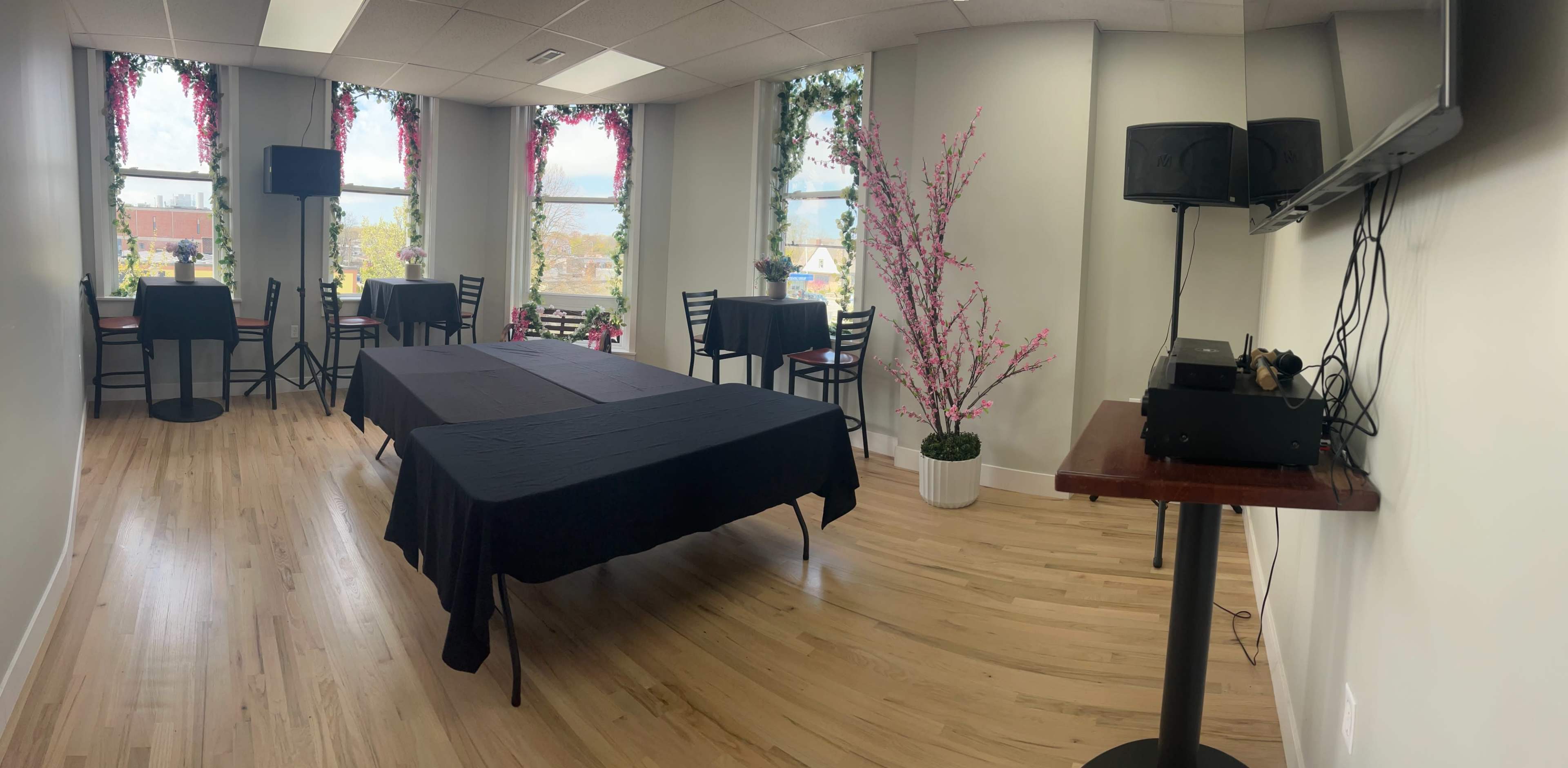 The image shows a spacious room with several tables covered in black tablecloths, surrounded by chairs, and decorated with floral arrangements and large windows letting in natural light.