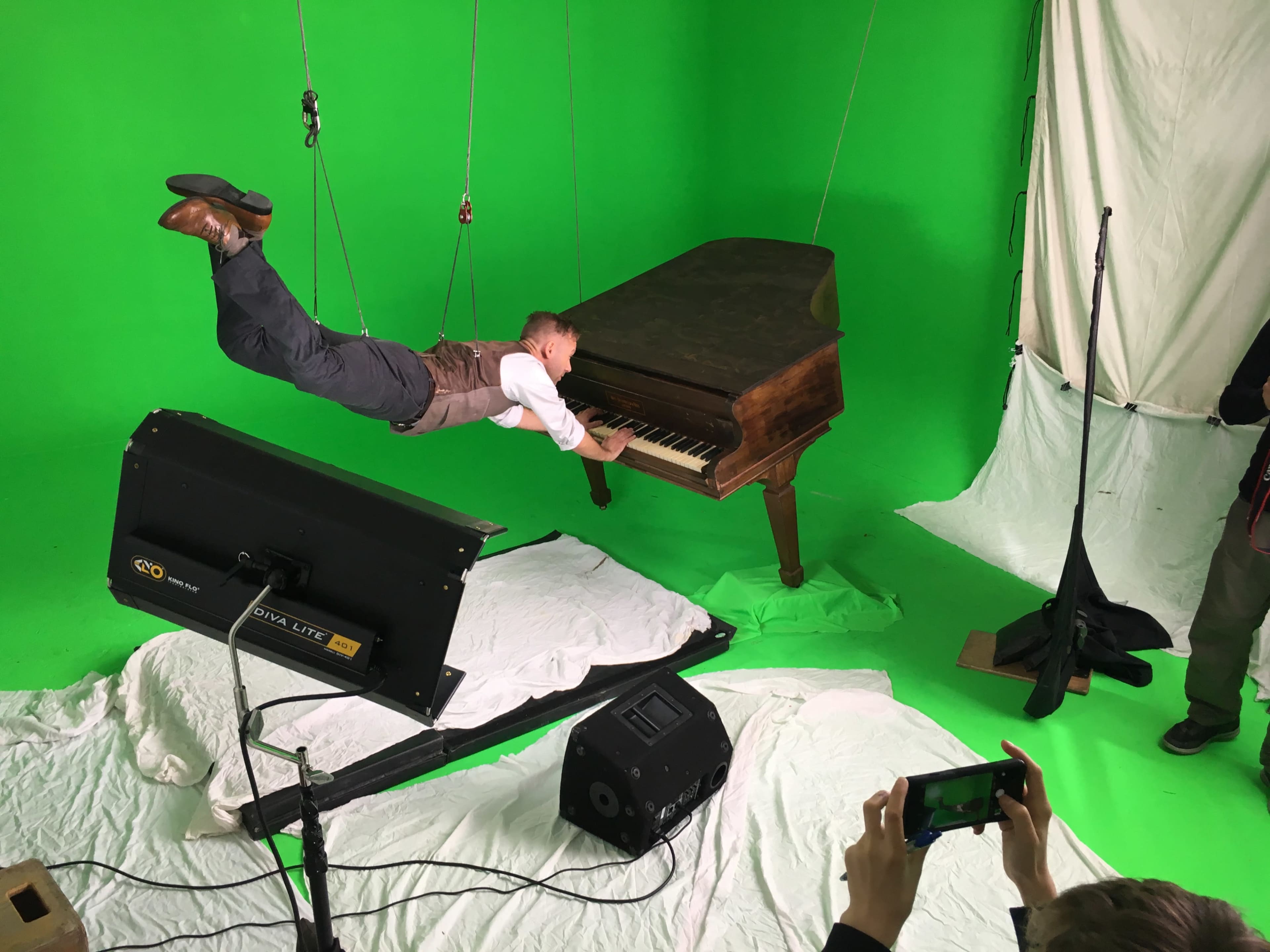 A person in a suit is suspended mid-air while playing a grand piano against a green screen backdrop.
