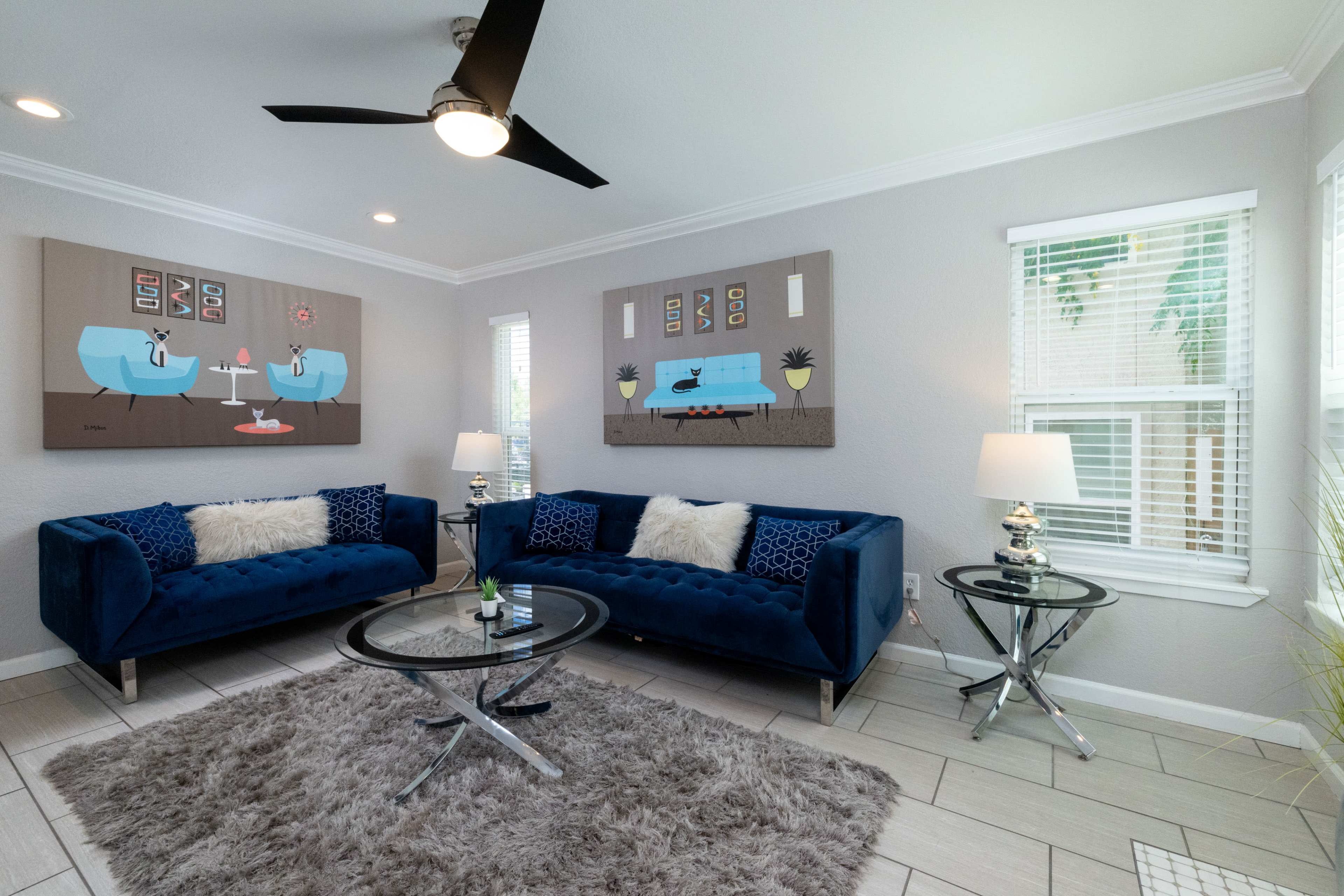 The image shows a modern living room with two blue velvet sofas, a glass coffee table, decorative lamps, and artwork on the walls.