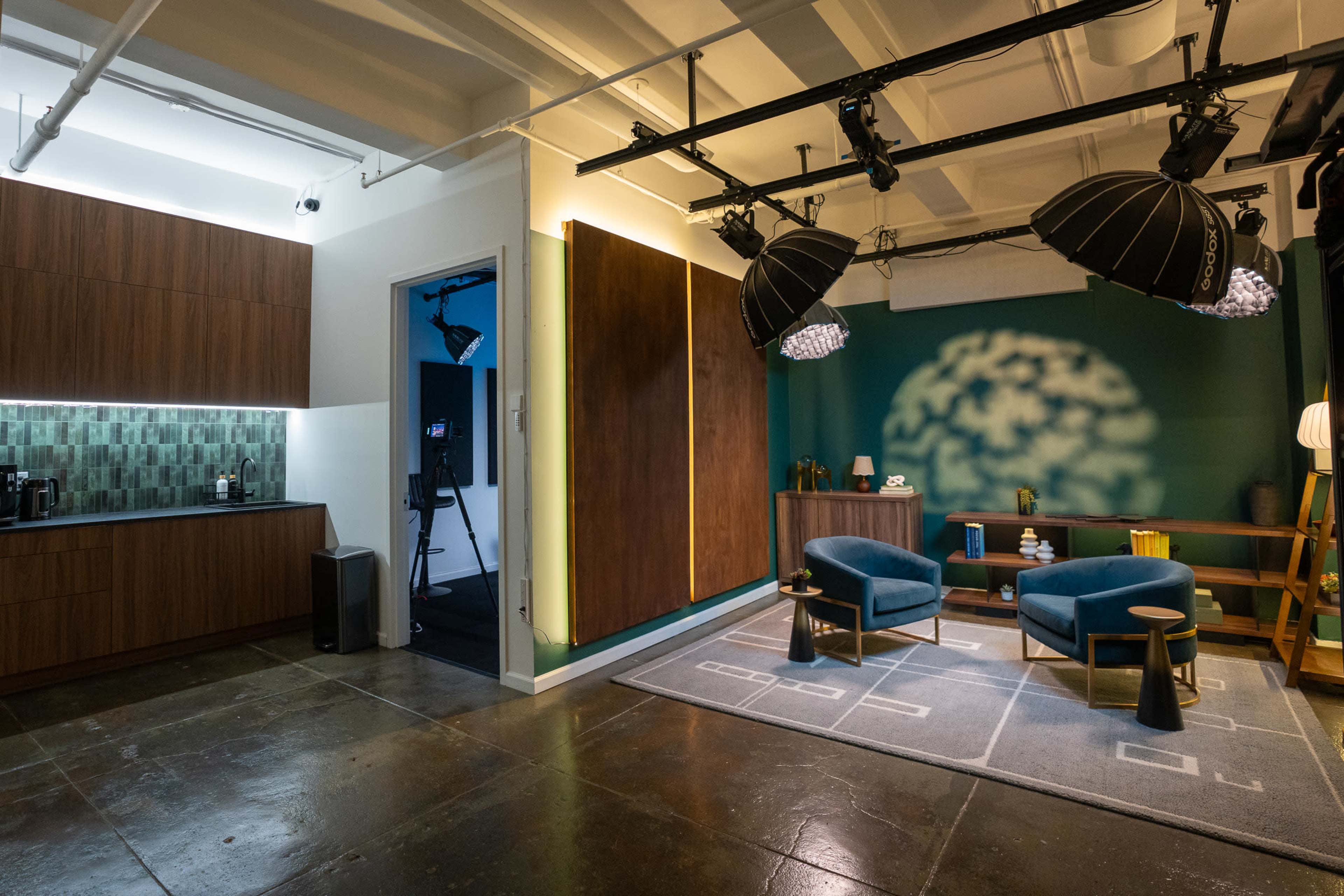 The image shows a modern studio space featuring a kitchenette area, a vibrant green wall with soft lighting, and two blue chairs arranged near a decorative wooden console table.