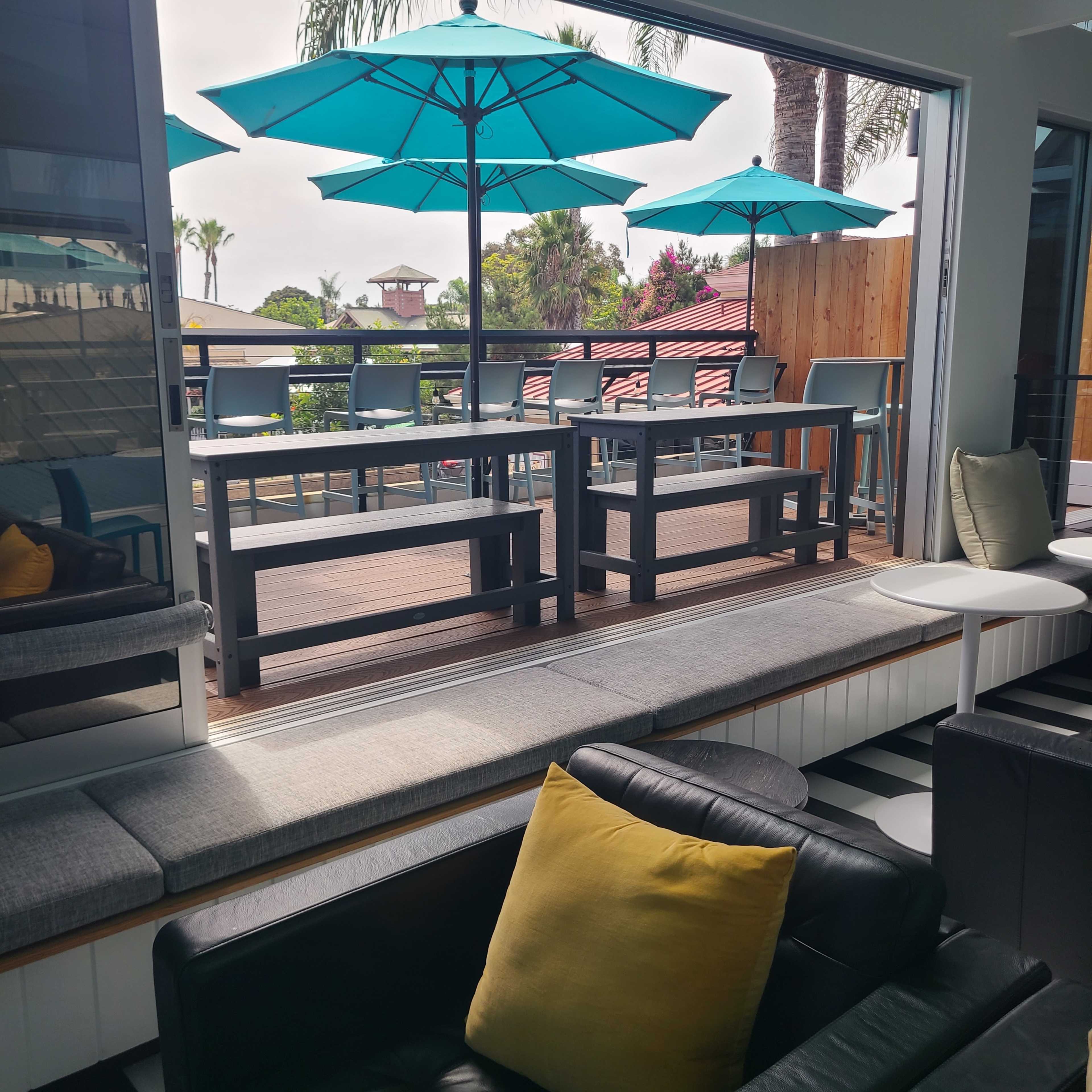 The image shows a modern indoor seating area with large windows looking out onto a deck featuring outdoor tables and teal umbrellas.
