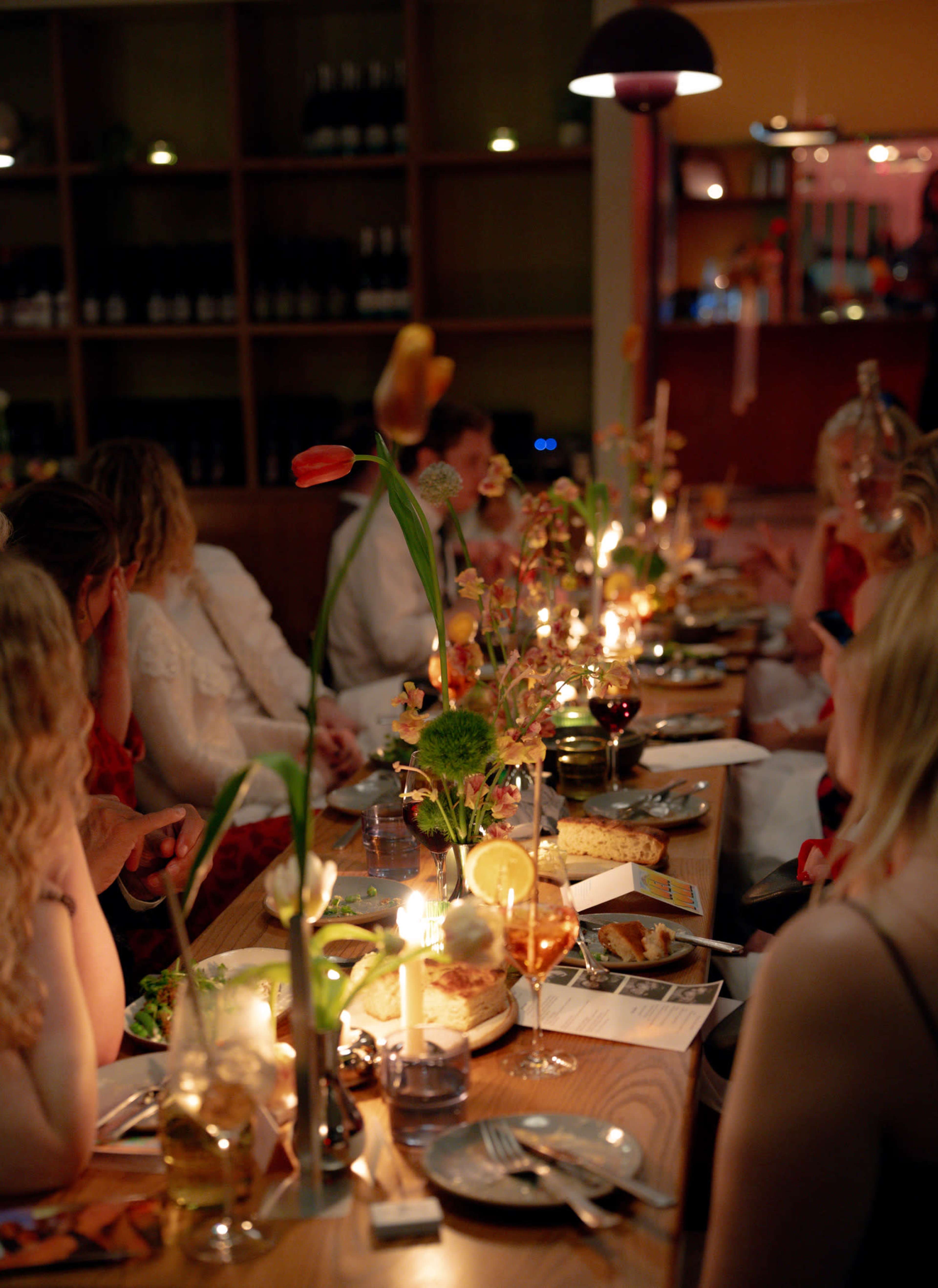 A long dining table is adorned with flowers and candles, with guests enjoying their meals in a warmly lit restaurant setting.