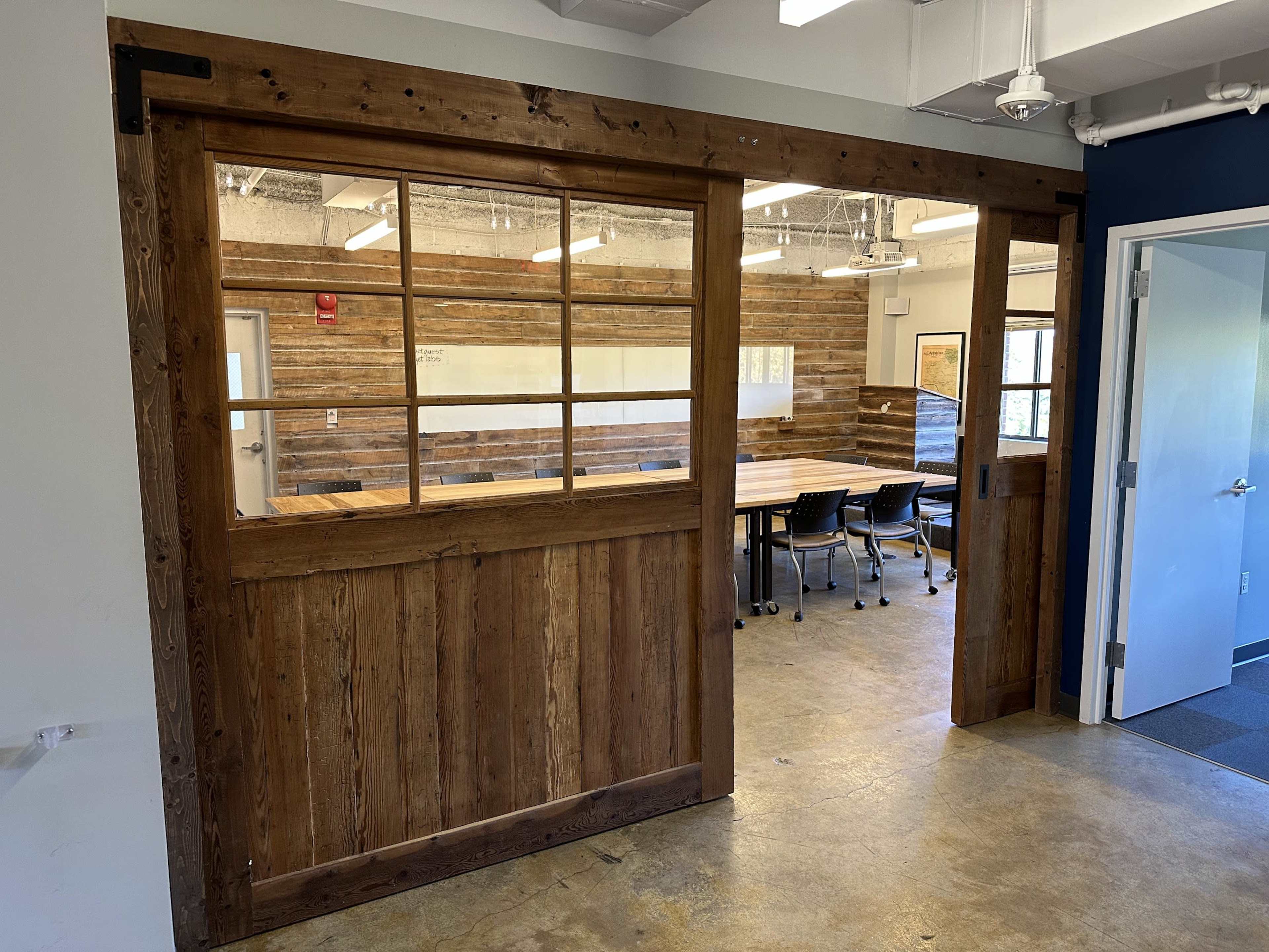 A wooden sliding door with windows opens to a meeting room featuring a long table and chairs against a rustic wooden wall.