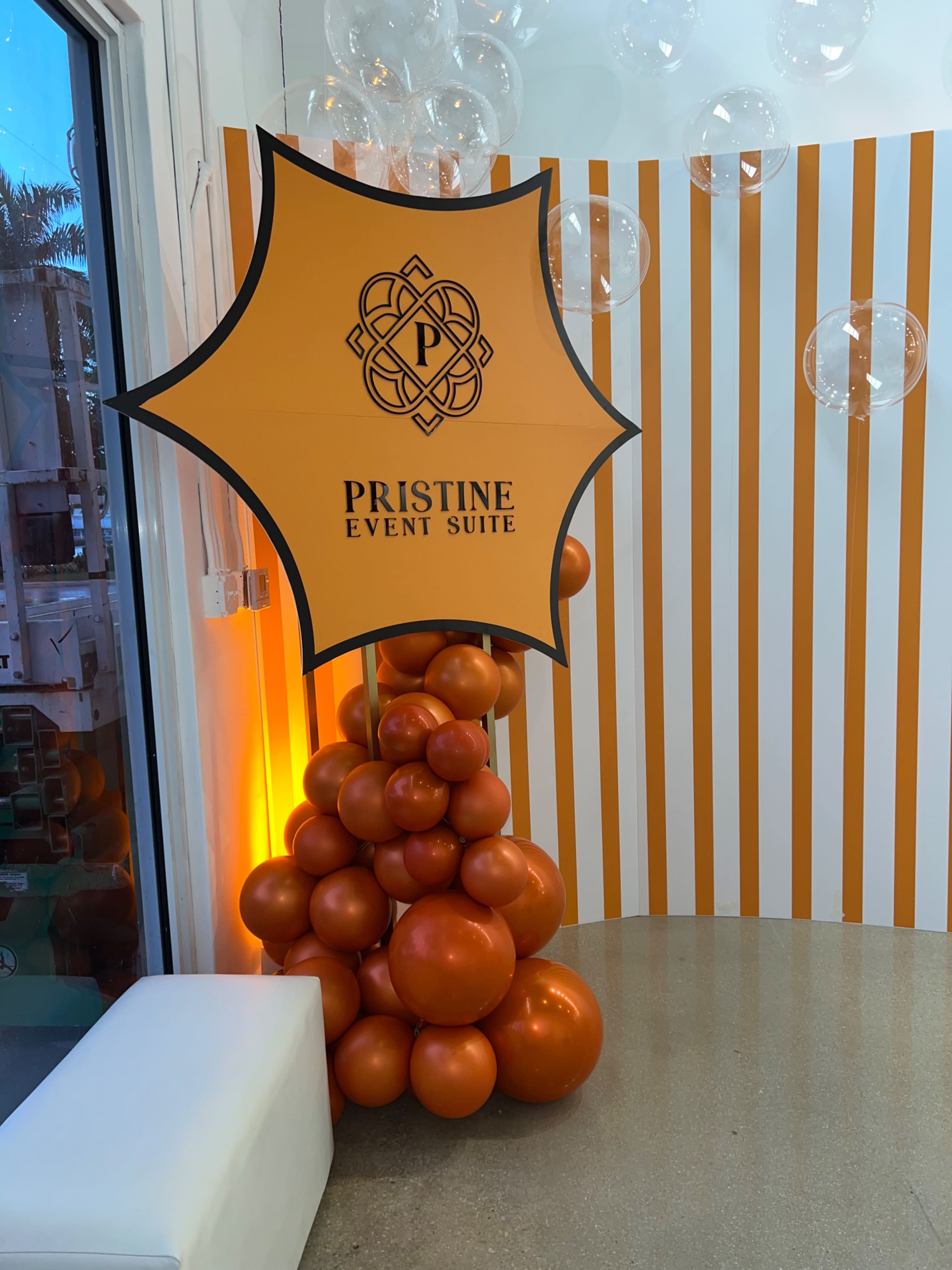 The image features a decorative display for "Pristine Event Suite," consisting of an orange and black sign surrounded by clusters of orange balloons against a striped background.
